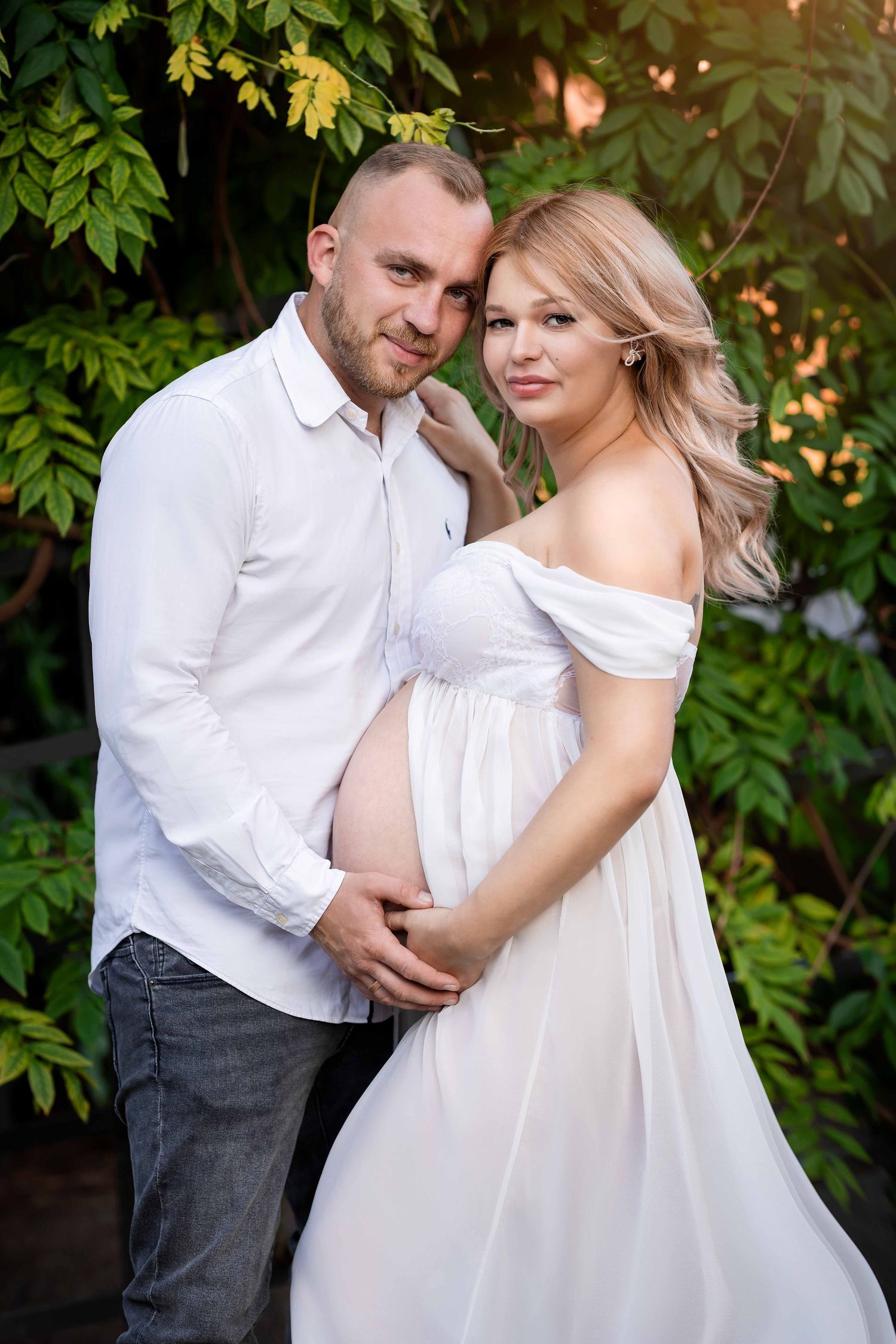 A romantic maternity photoshoot in a garden setting with couple wearing white and embracing tenderly.