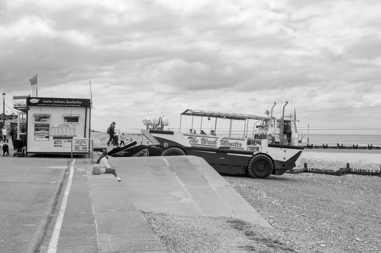 Panoramic view of a beach parking lot with cars and buildings under a cloudy sky along the coastline.