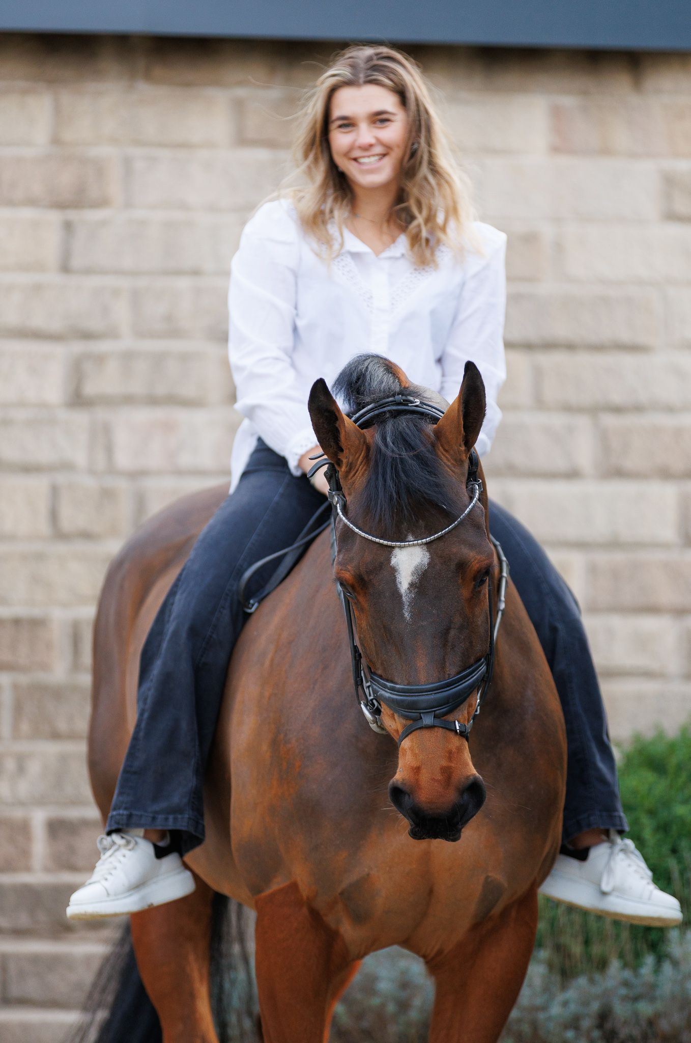 A beautiful bay horse wearing a black bridle stands next to someone in a white lace dress in an outdoor setting.
