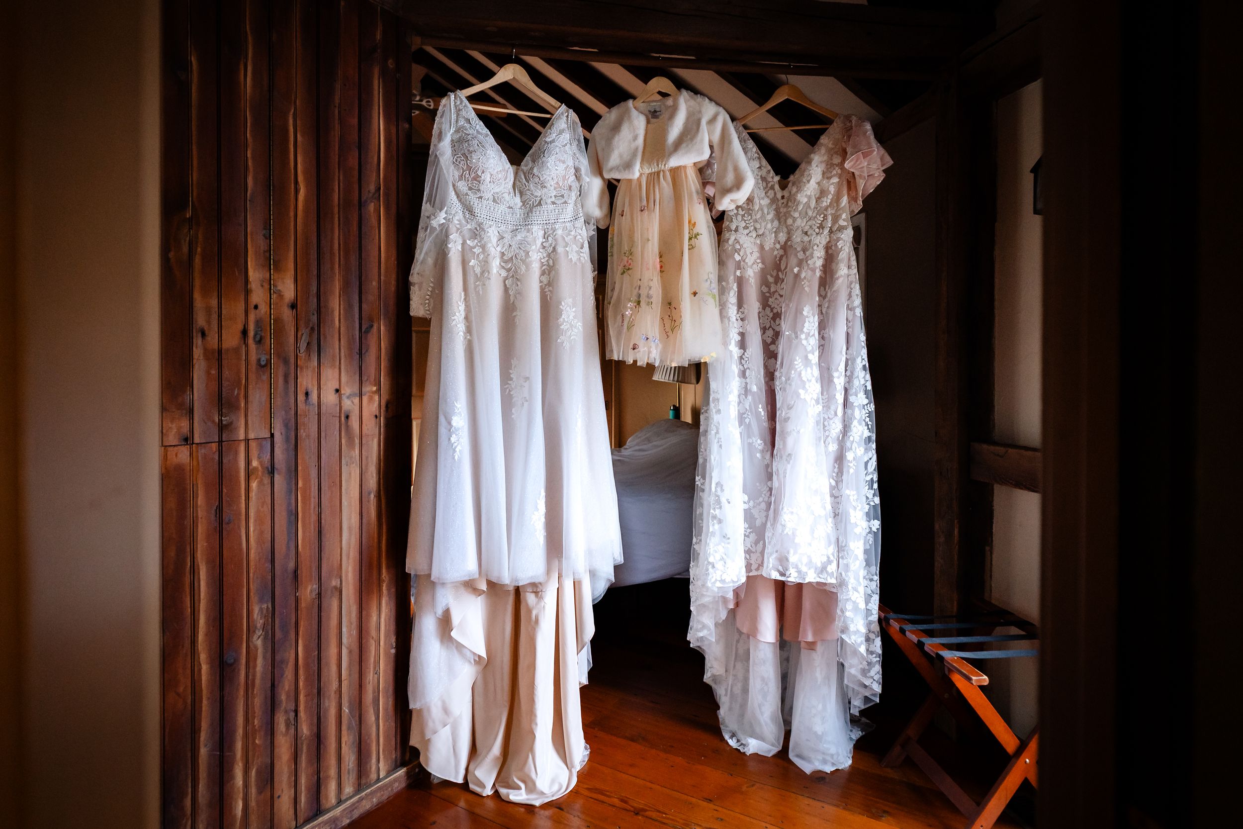 Vintage-style white lace dresses and gowns hanging on wooden hangers in a rustic room with natural lighting.