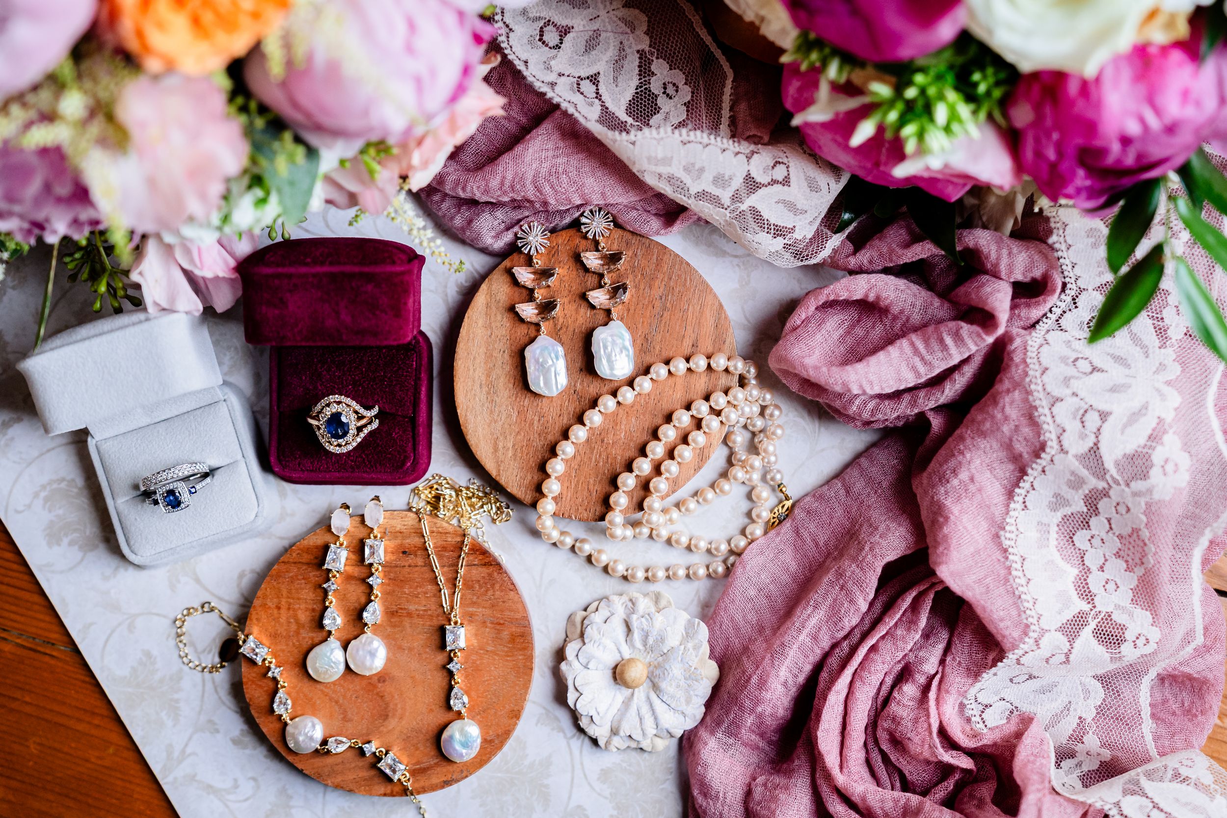 Flatlay of wedding jewelry including pearl necklaces, rings in burgundy box, and pink fabric on wooden surface.