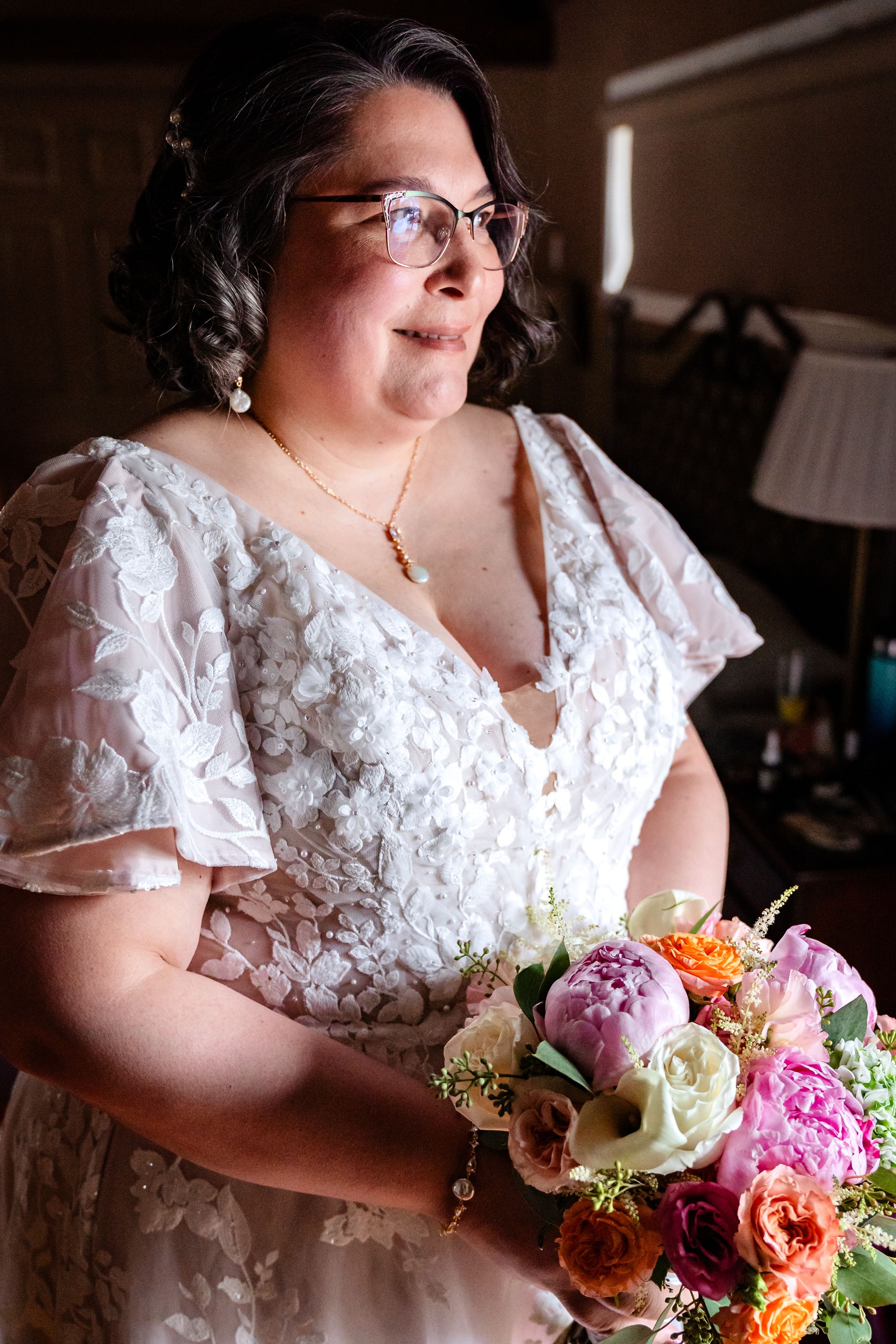 Close-up view of a bride in a lace dress holding a vibrant bouquet with pink, orange and white flowers.