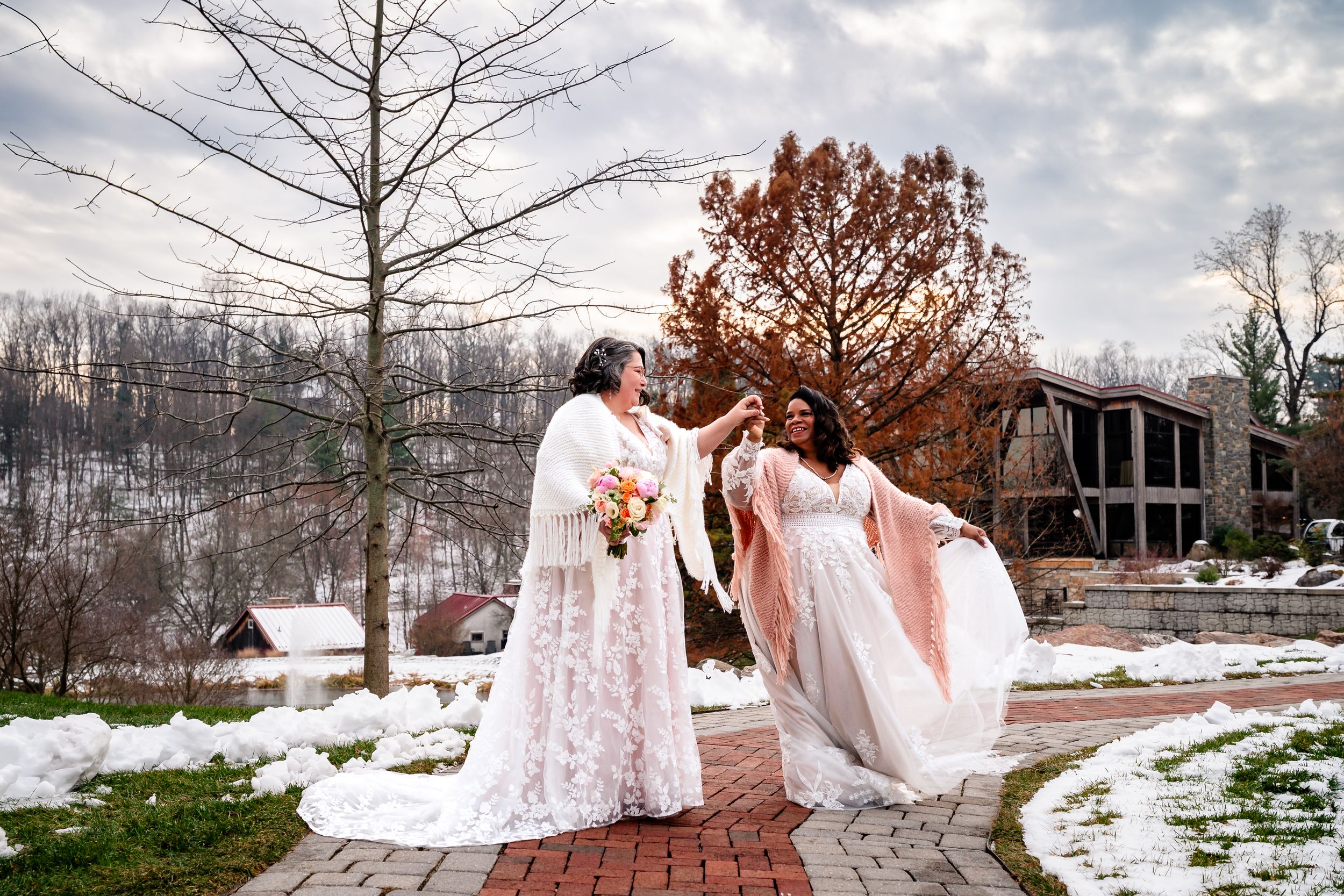 Wedding party in white dresses poses together in a snowy winter landscape with rustic barn backdrop.