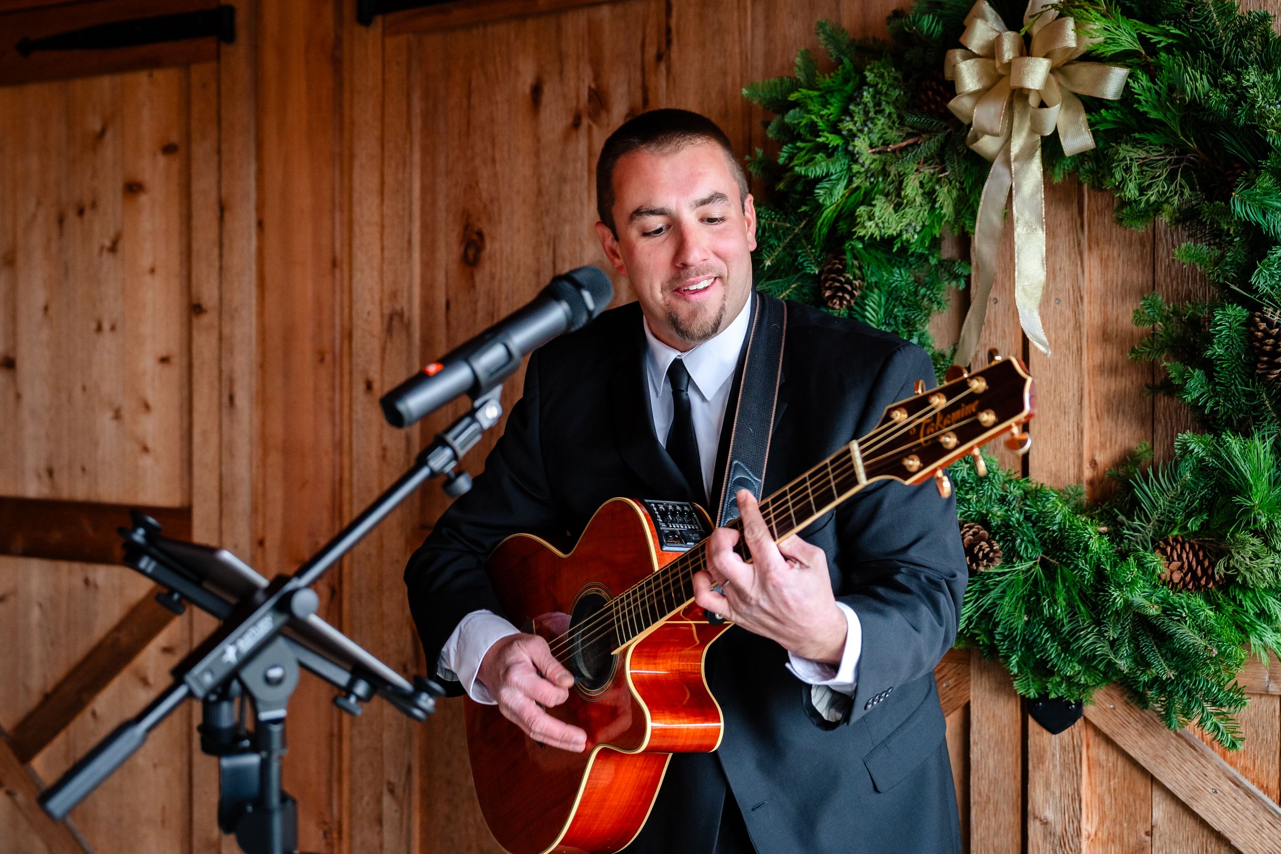 Musician performs with acoustic guitar inside rustic wooden barn decorated with holiday greenery.