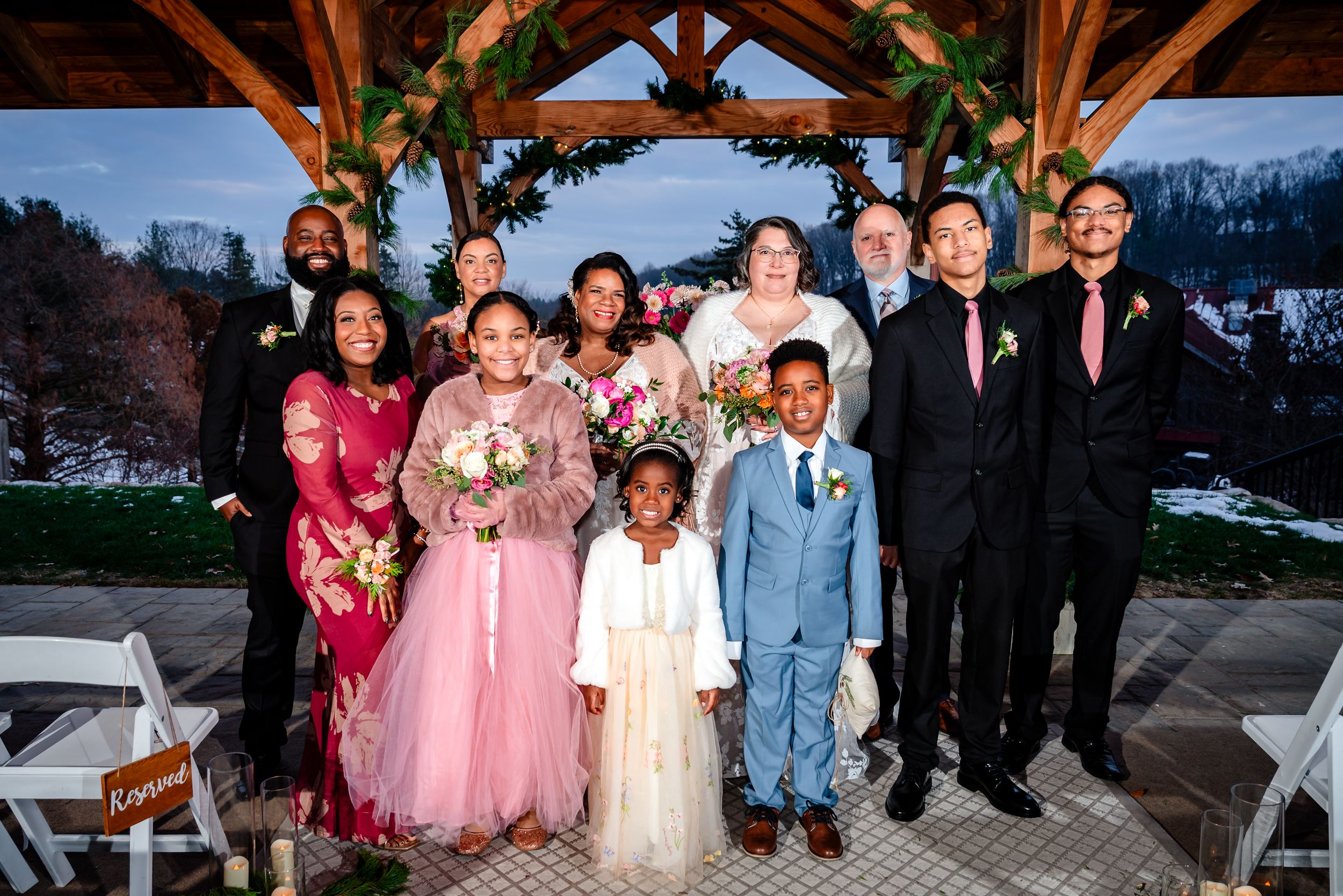 Wedding party group photo outdoors at a rustic wooden pavilion with snow-covered mountains in background.
