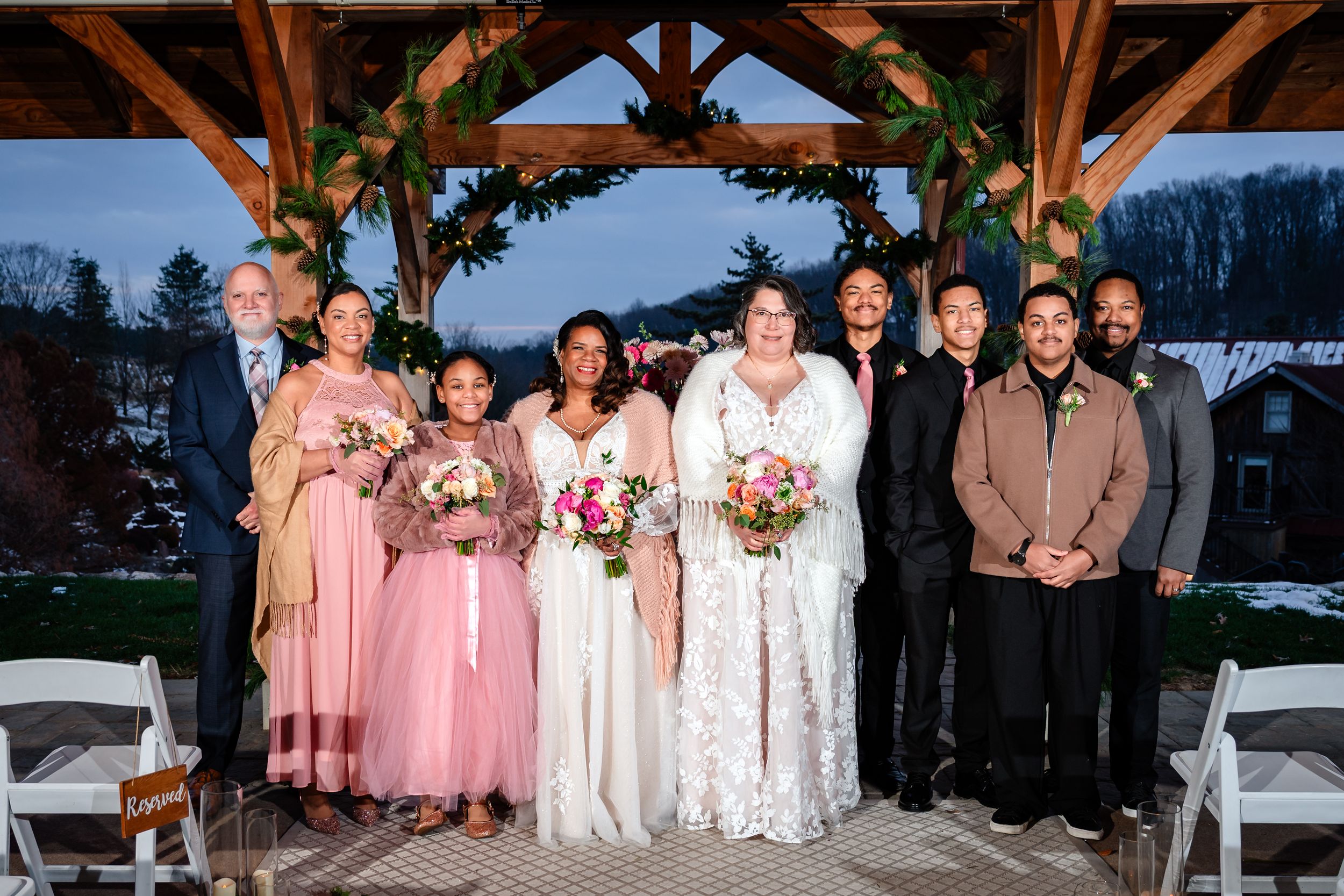 Wedding party in pink dresses and suits posing under a wooden pavilion at dusk.