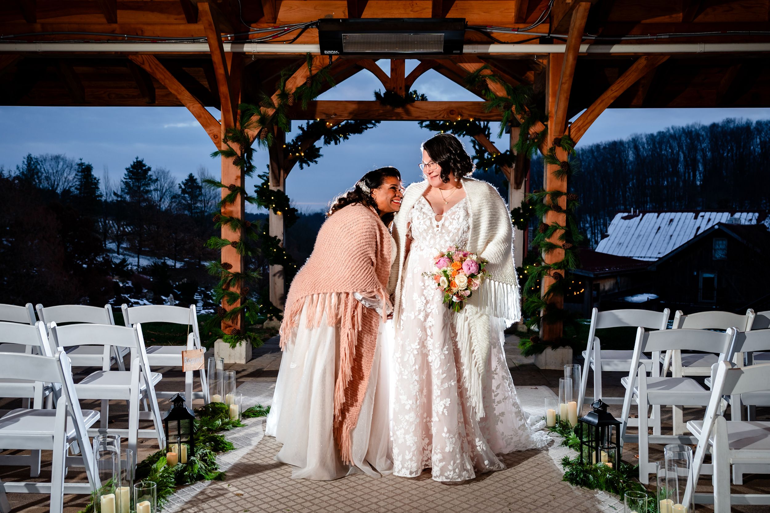 Wedding ceremony setup with white chairs under a wooden pavilion against mountain backdrop at sunset.