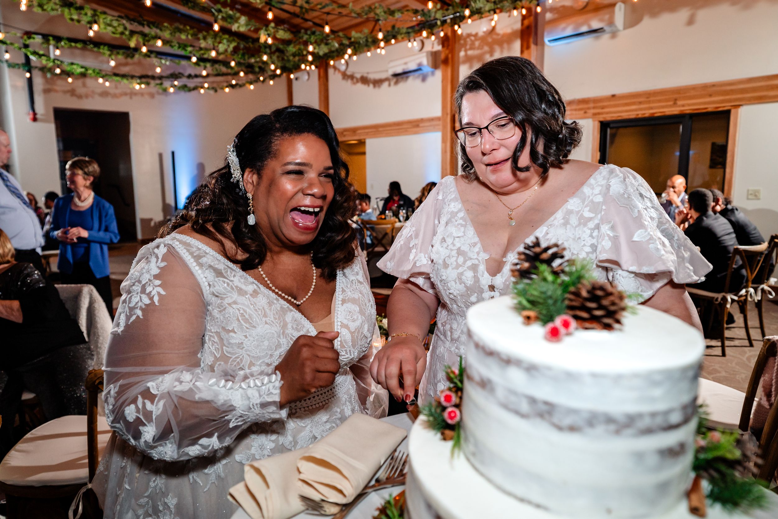 Two people in white lace dresses share a joyful moment while cutting their wedding cake at an indoor reception.