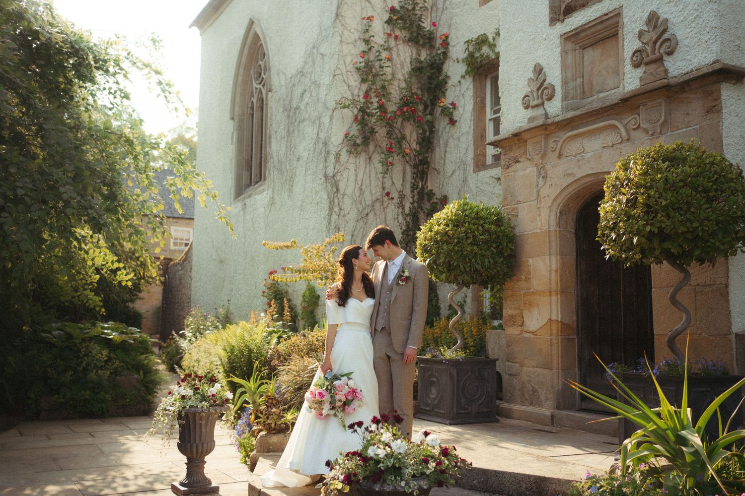 A couple in formal wedding attire walks through a sunlit garden path beside a historic stone building.