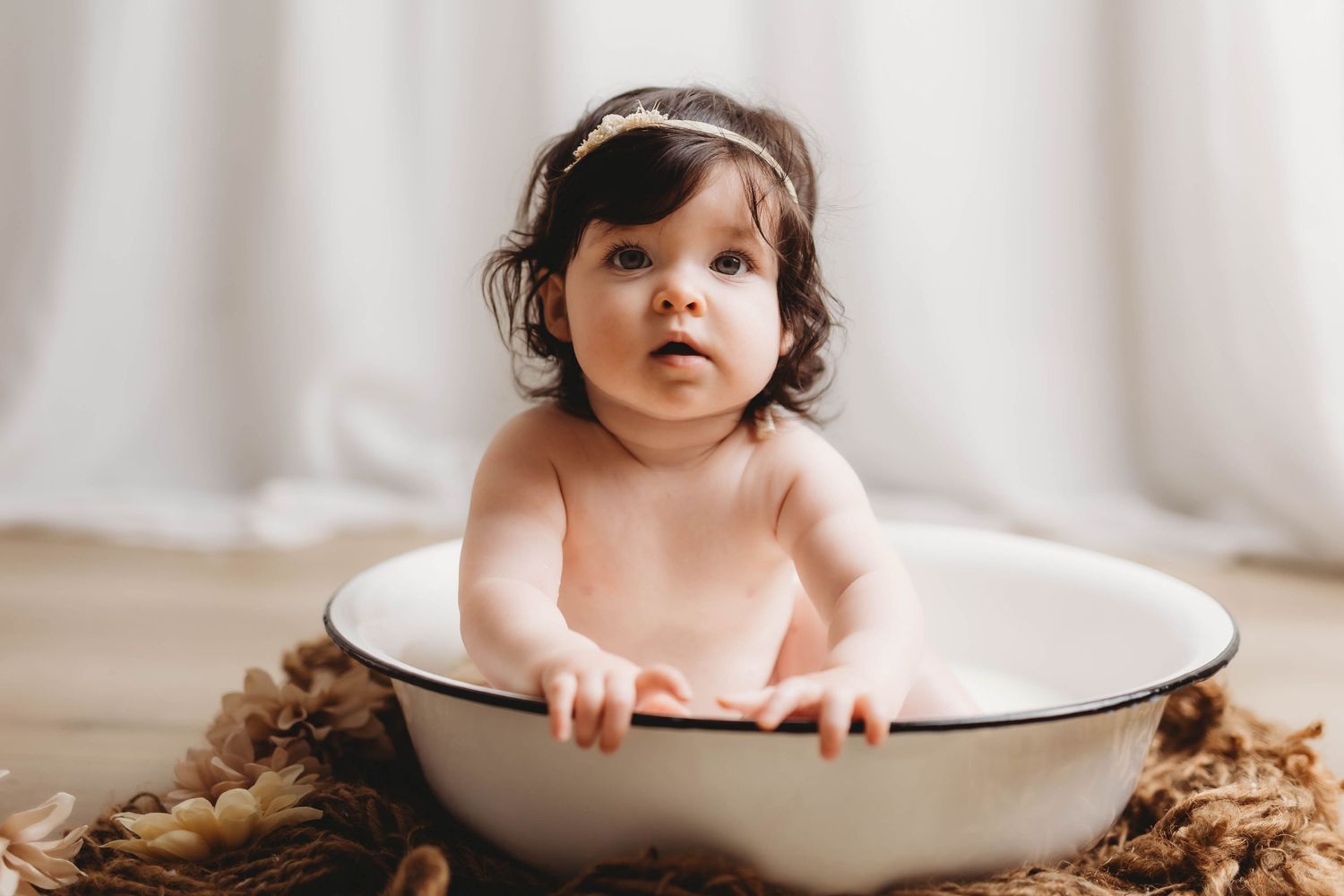 Baby sitting in white enamel basin surrounded by cozy brown blankets during milk bath photo session.