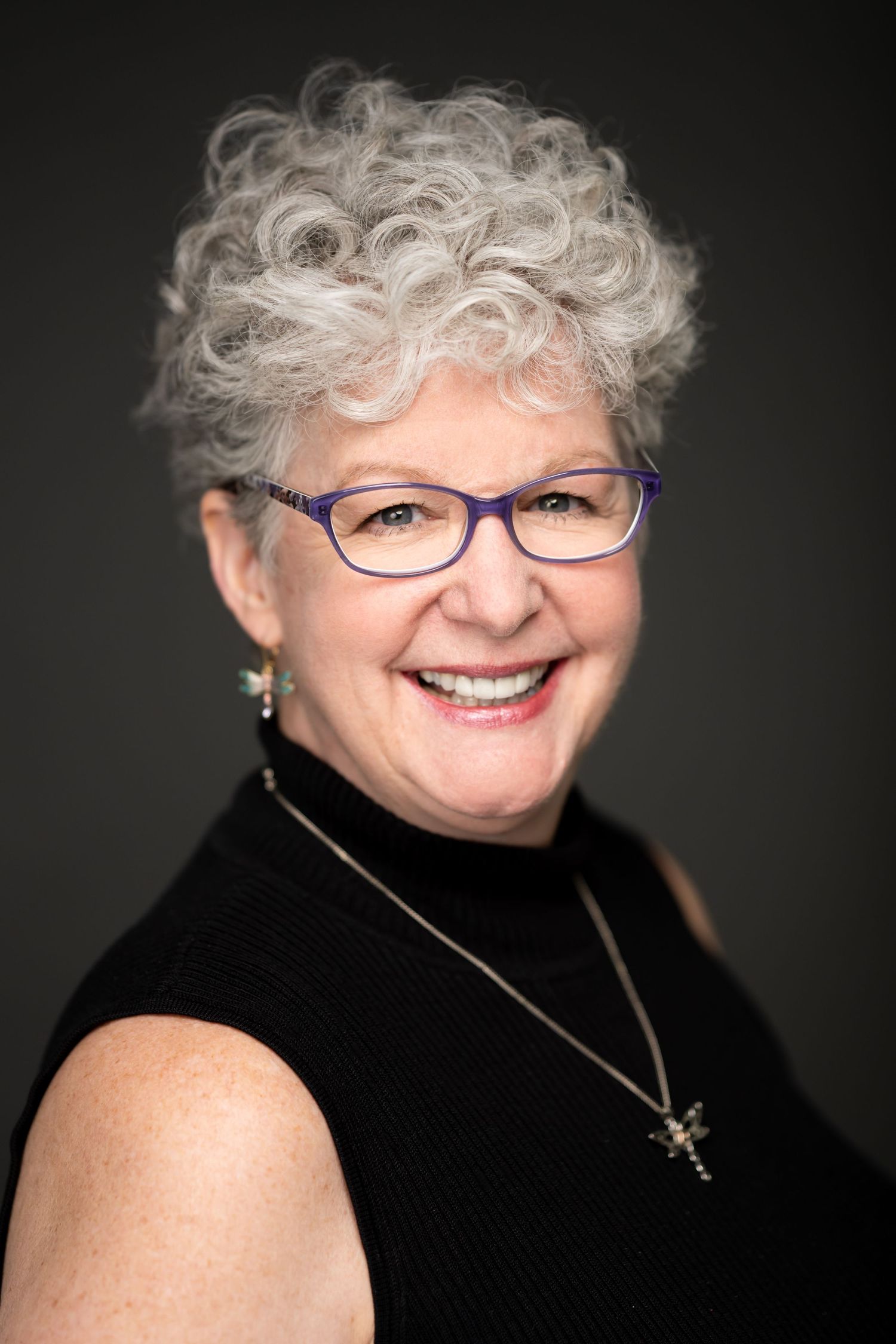 Portrait of person with silver curly hair wearing glasses and black turtleneck against dark background.