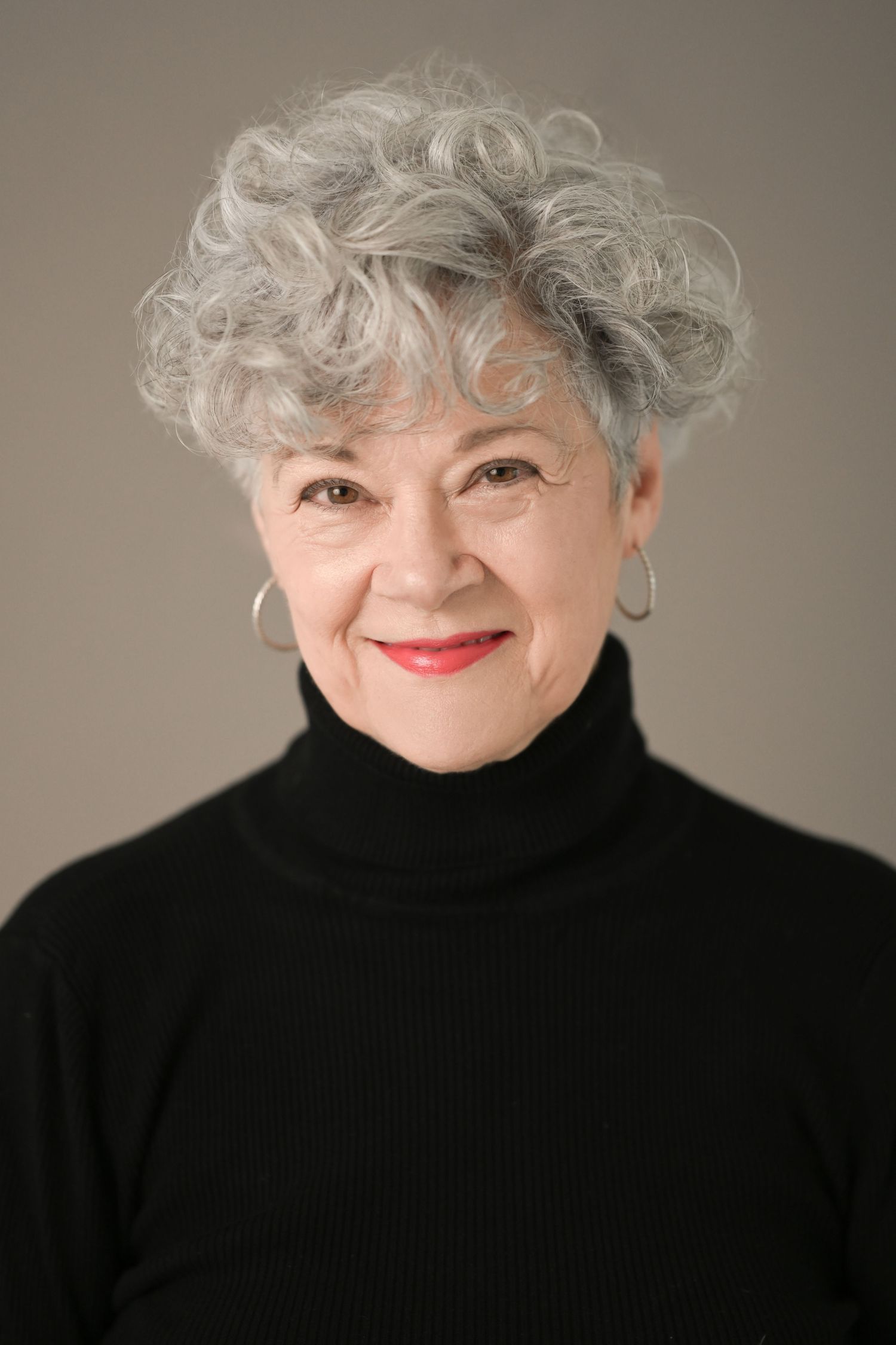 Close-up profile shot of person with silver curly hair wearing black turtleneck against neutral background.