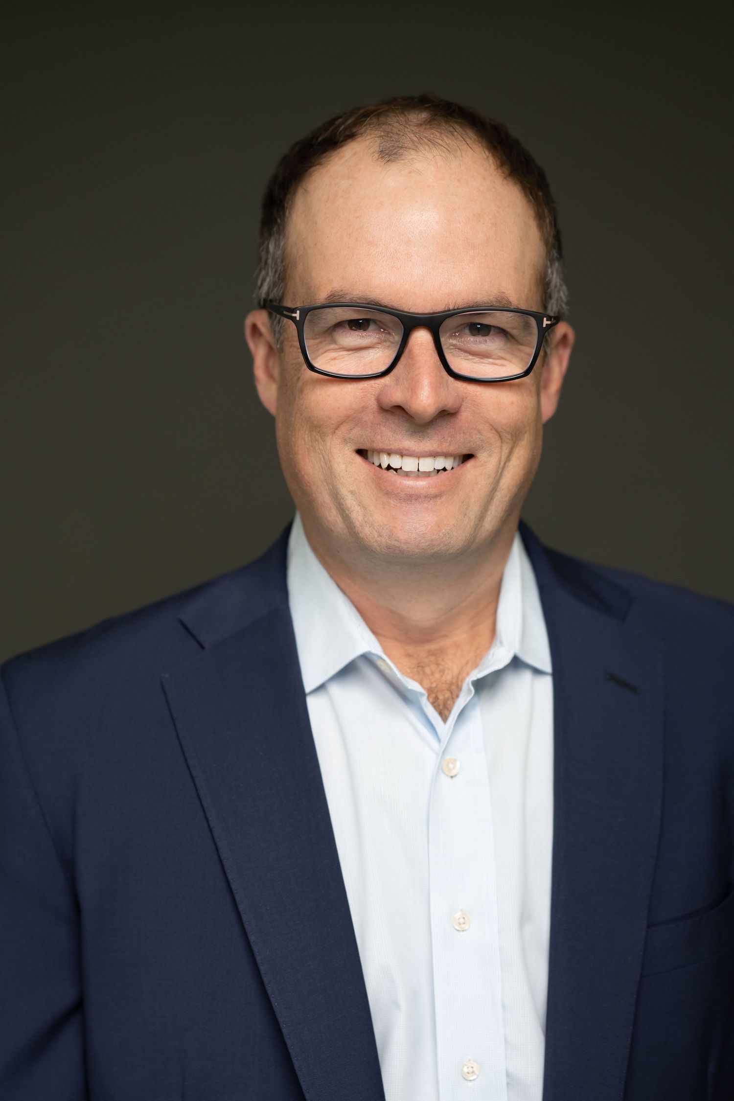 Professional headshot of person in navy suit and glasses against dark background.