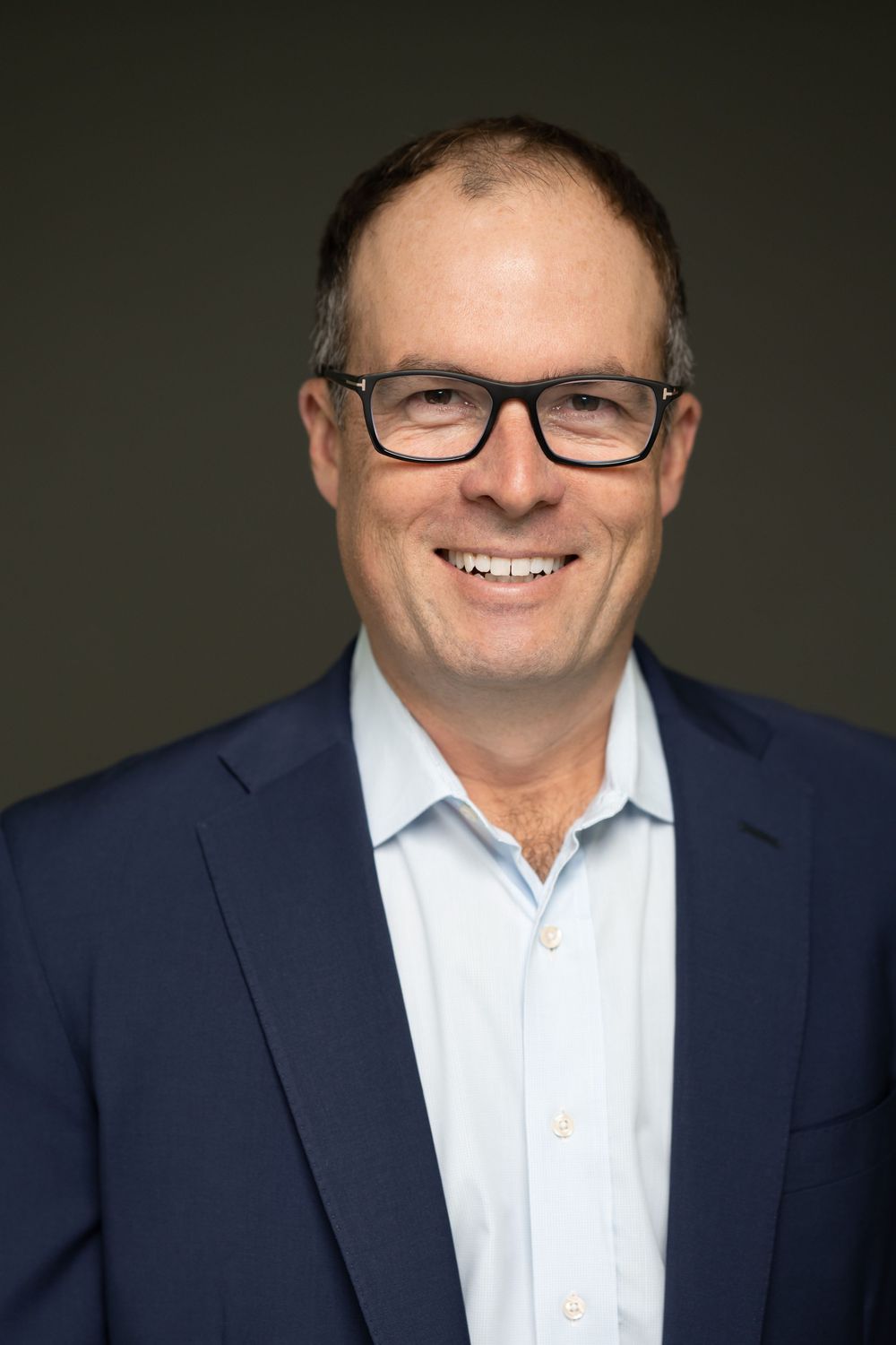 Professional headshot of person in navy suit and glasses against dark background.