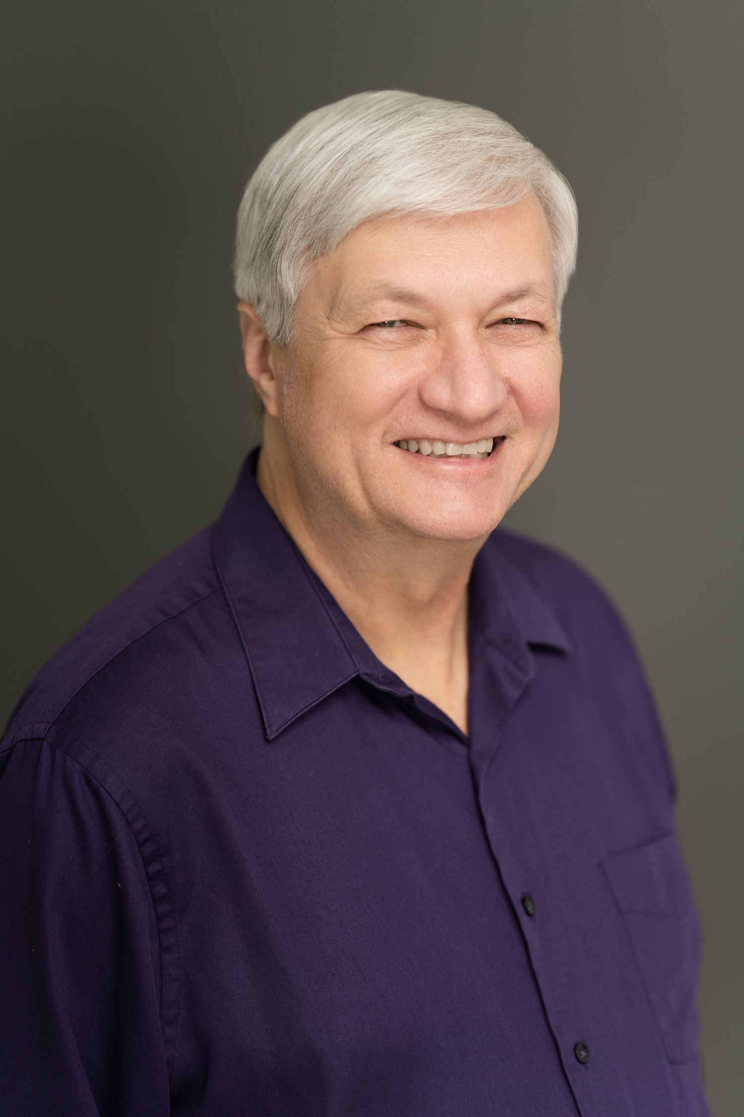 Professional headshot of person in navy blue shirt against gray background showing joyful expression.