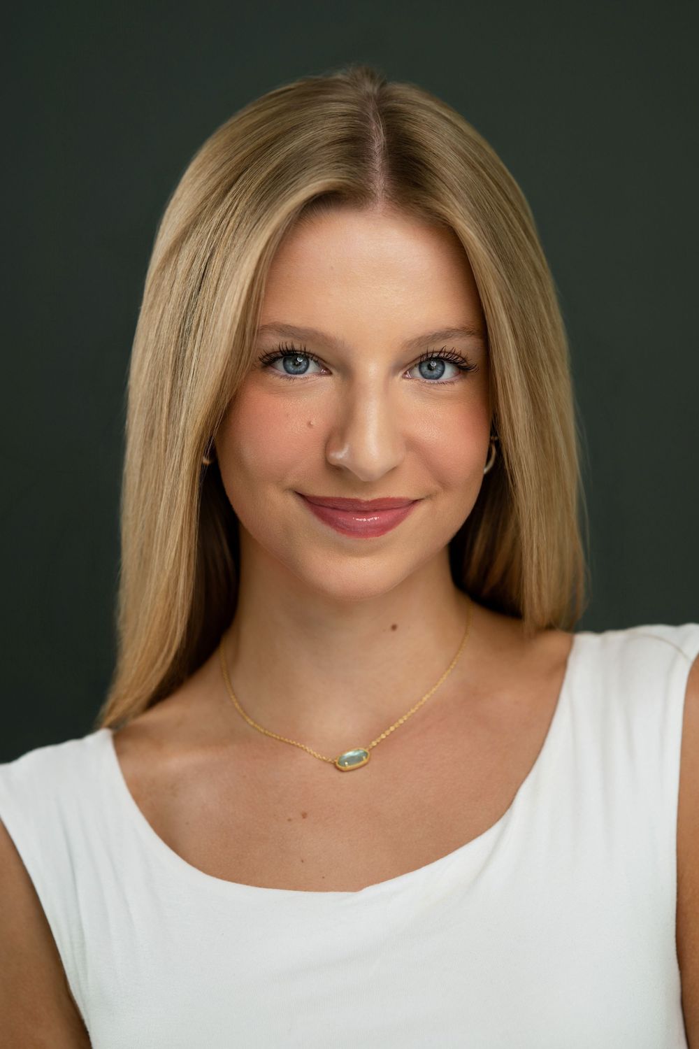Young blonde woman with blue eyes wearing a white top and gold necklace, smiling in a professional headshot.
