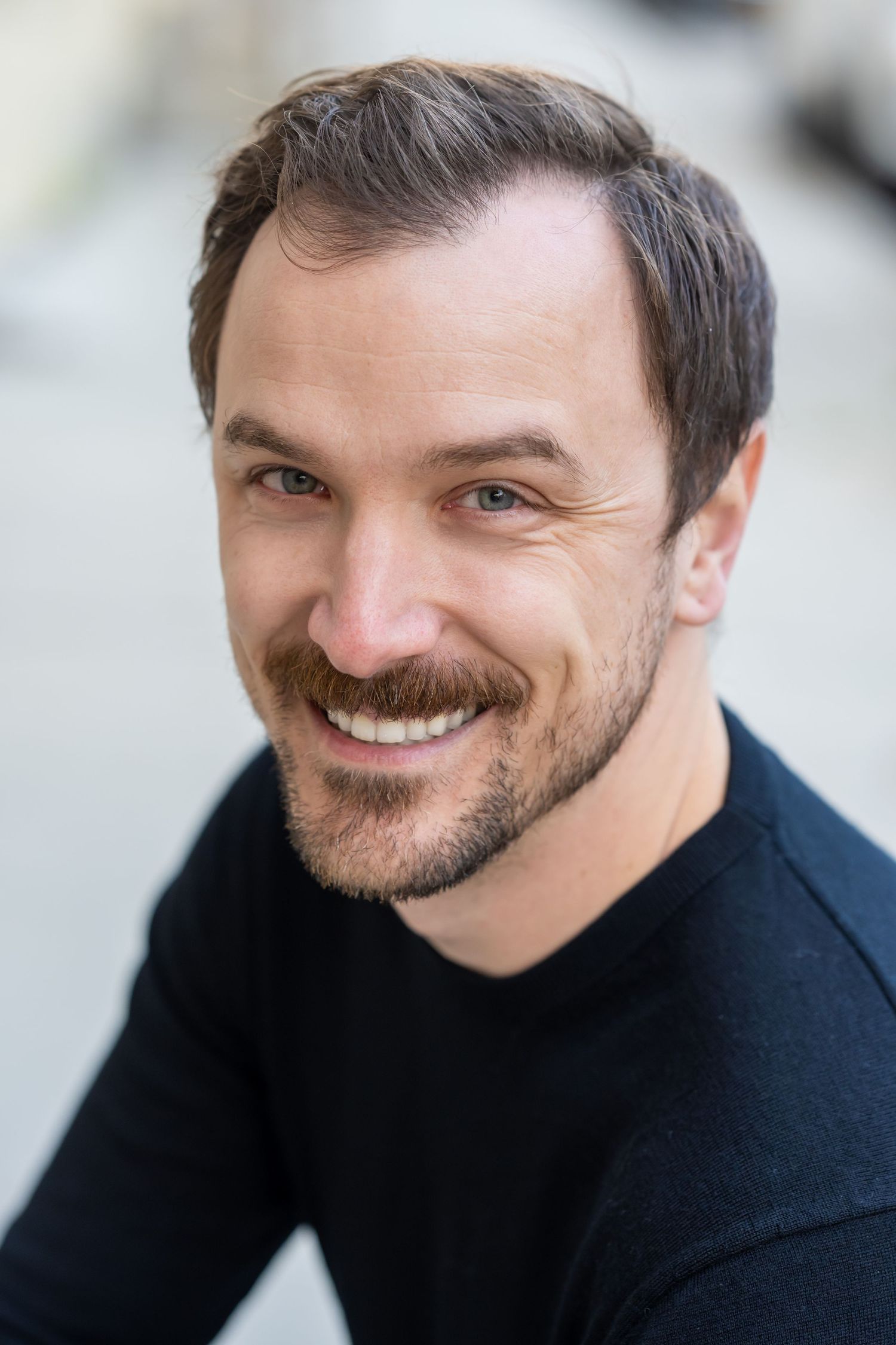 Close-up portrait of person with dark facial hair wearing black shirt against light background.