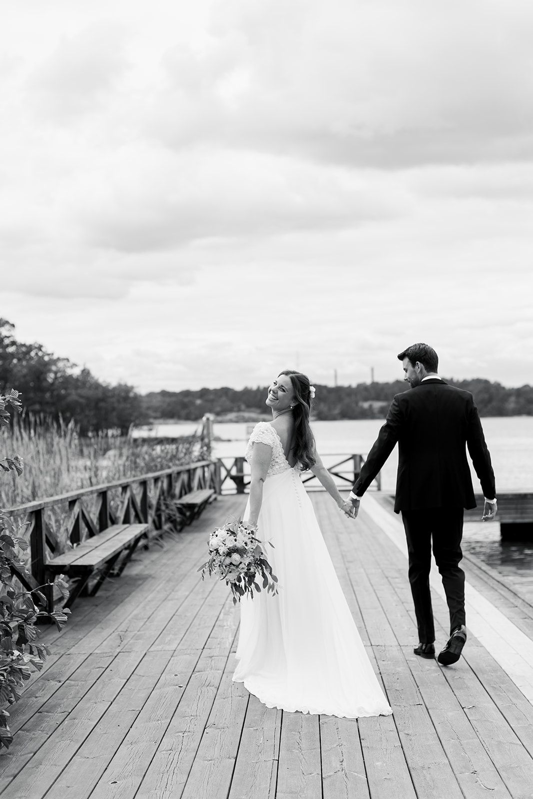 A bride and groom walk hand in hand along a wooden dock by a lake on their wedding day in black and white.