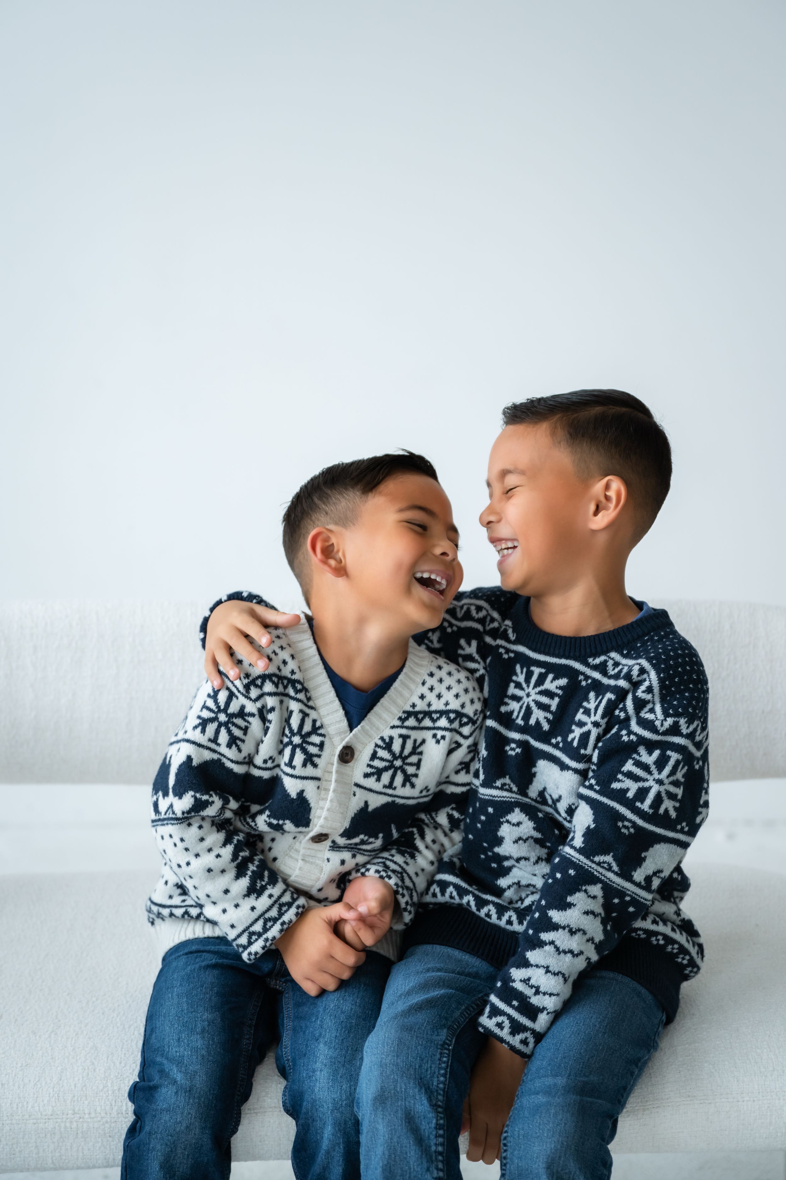 Two children wearing matching blue and white snowflake sweaters share playful moments on a white couch.