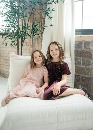Two children in velvet dresses sit together on a white couch with a potted bamboo plant and exposed brick wall behind them.
