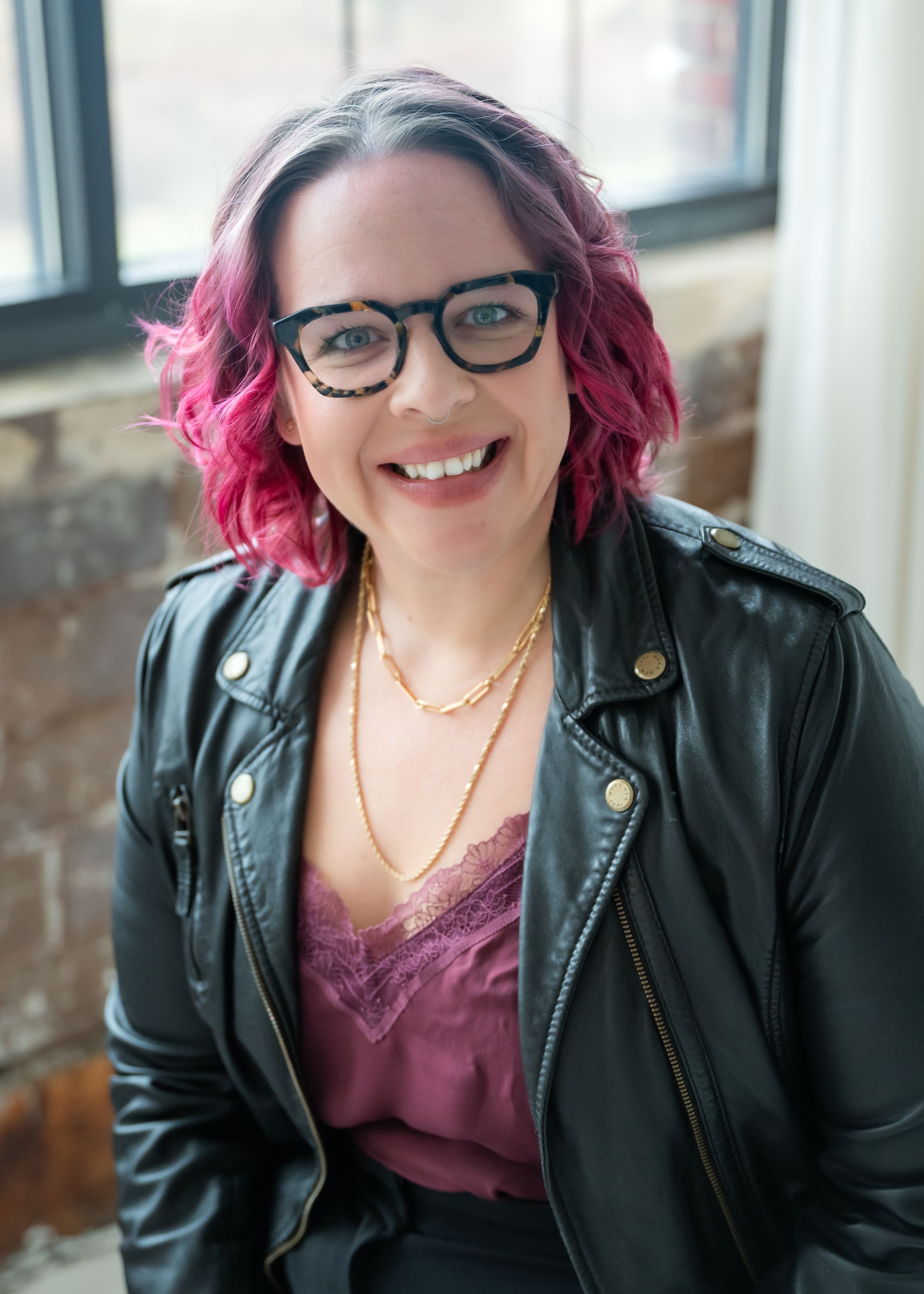 Woman with vibrant pink hair and glasses wearing a black leather jacket and purple top smiles brightly.