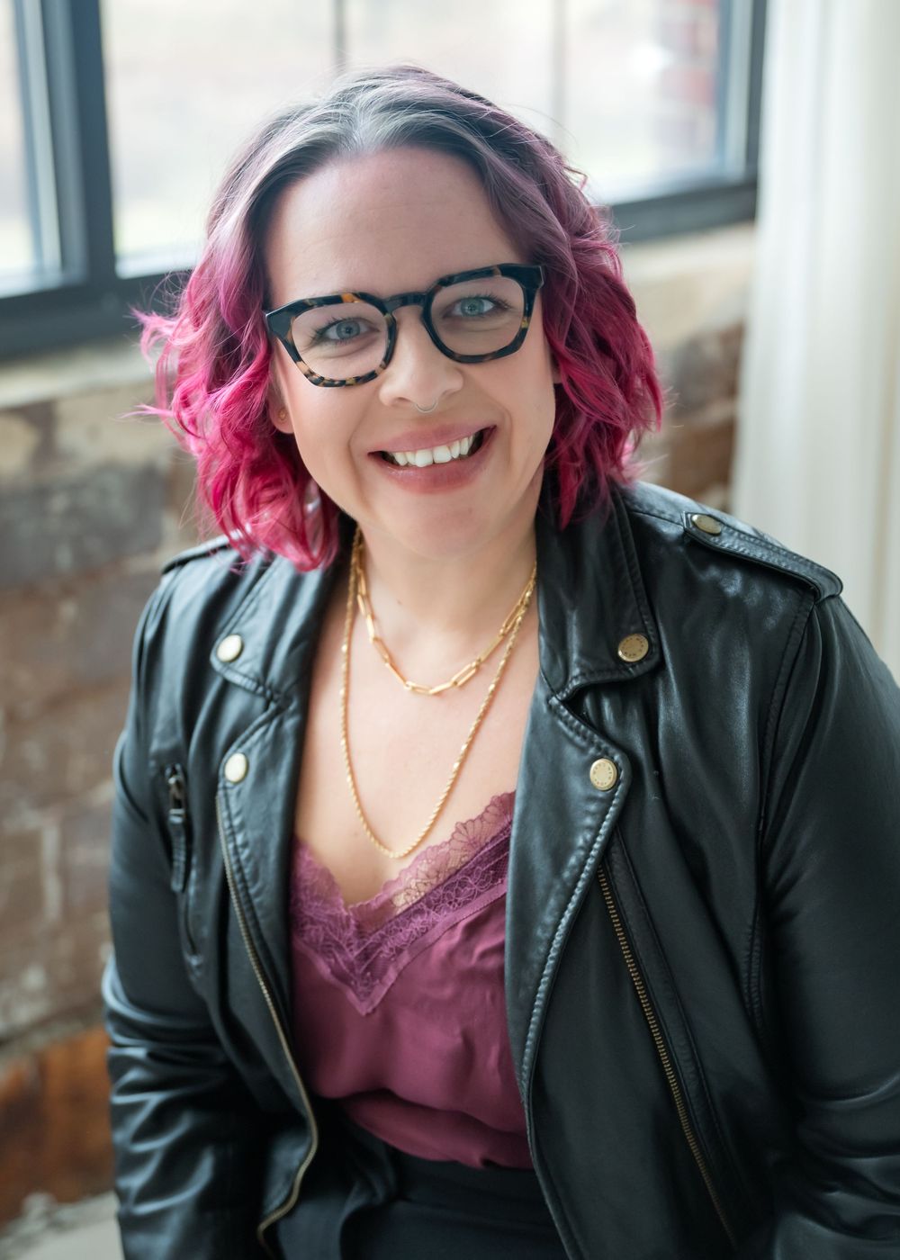 Woman with vibrant pink hair and glasses wearing a black leather jacket and purple top smiles brightly.