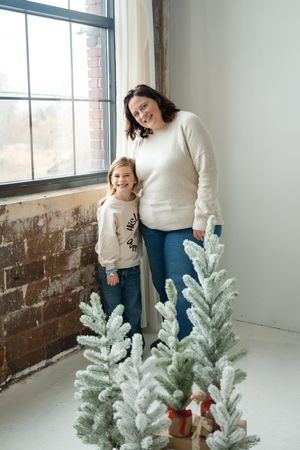 Mother and daughter decorate a frosted white Christmas tree together in a bright industrial-style room.