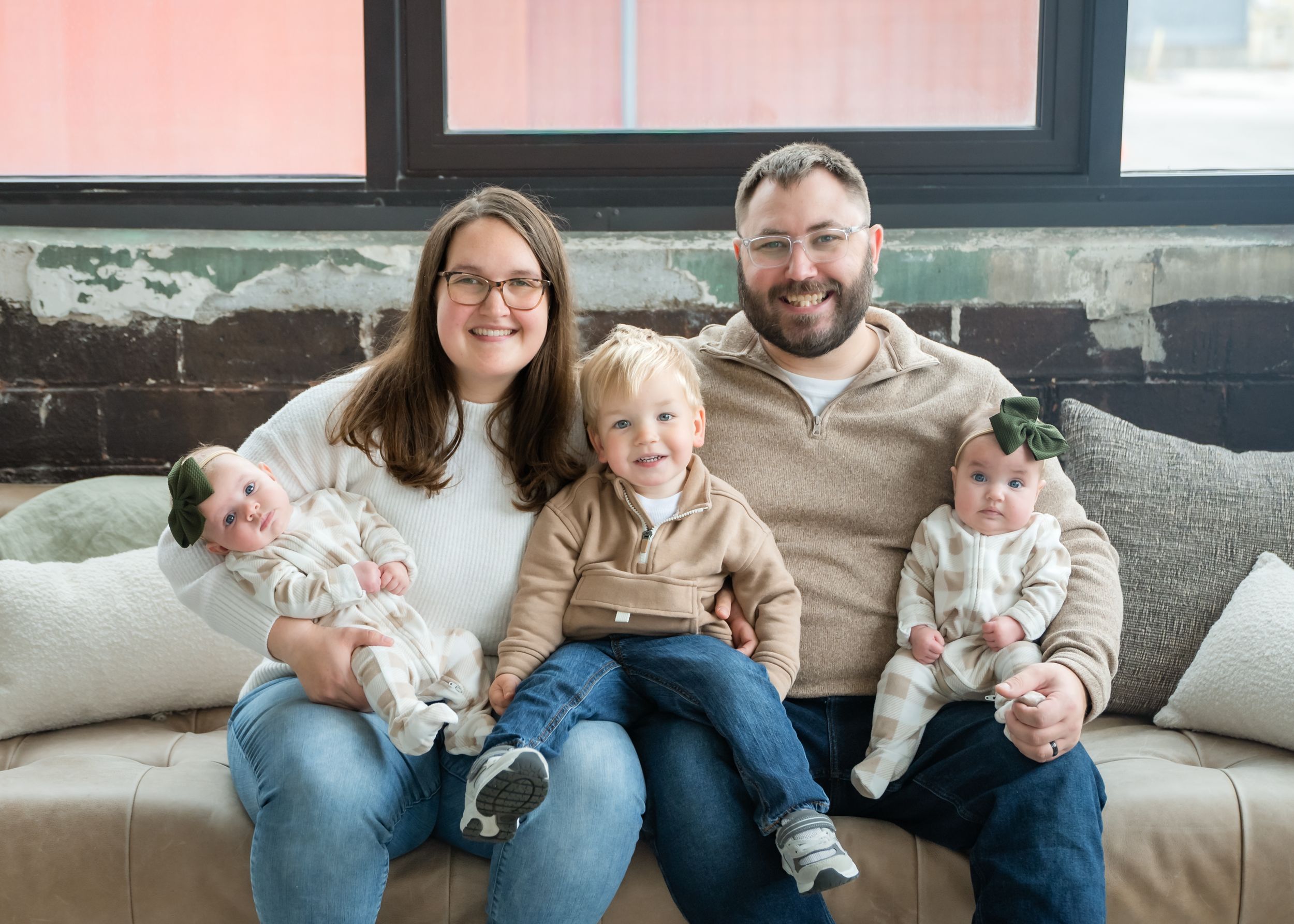 Family sits together on gray couch sharing tender moments with baby and toddler.