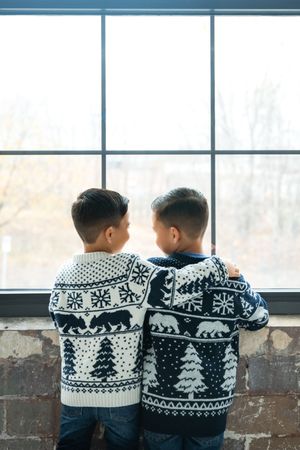 Two children wearing matching black and white Nordic pattern sweaters with bears and trees sit together by a window.