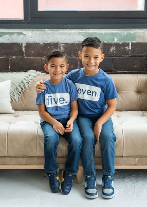 Two children in matching blue shirts and jeans sit together on a beige leather couch.