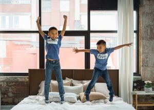 Two energetic children jumping playfully on a white bed near large windows in a modern room.