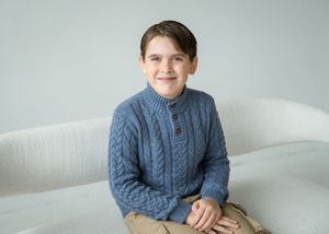 Young boy wearing a blue cable knit sweater sits on a white couch.