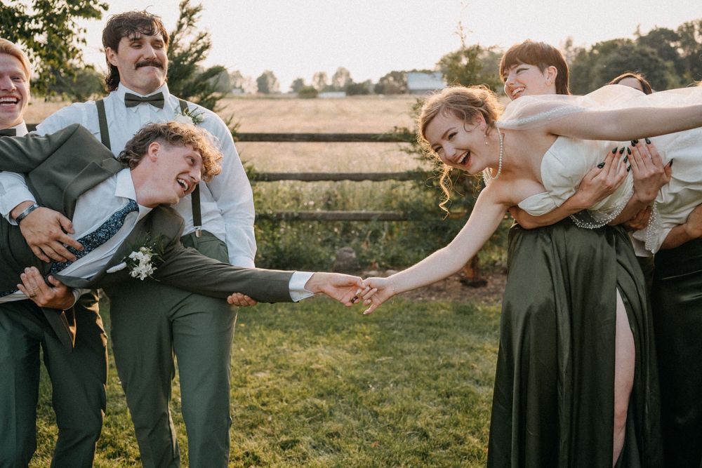 Wedding party members in formal attire playfully pose together on grassy field at sunset.