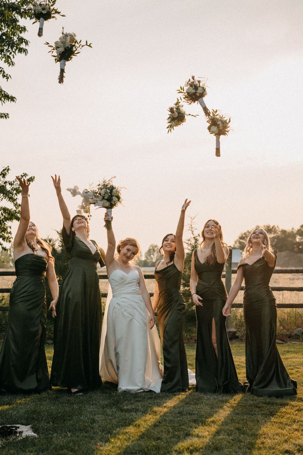 Wedding party in black dresses and white gown pose together on grass during sunset celebration.