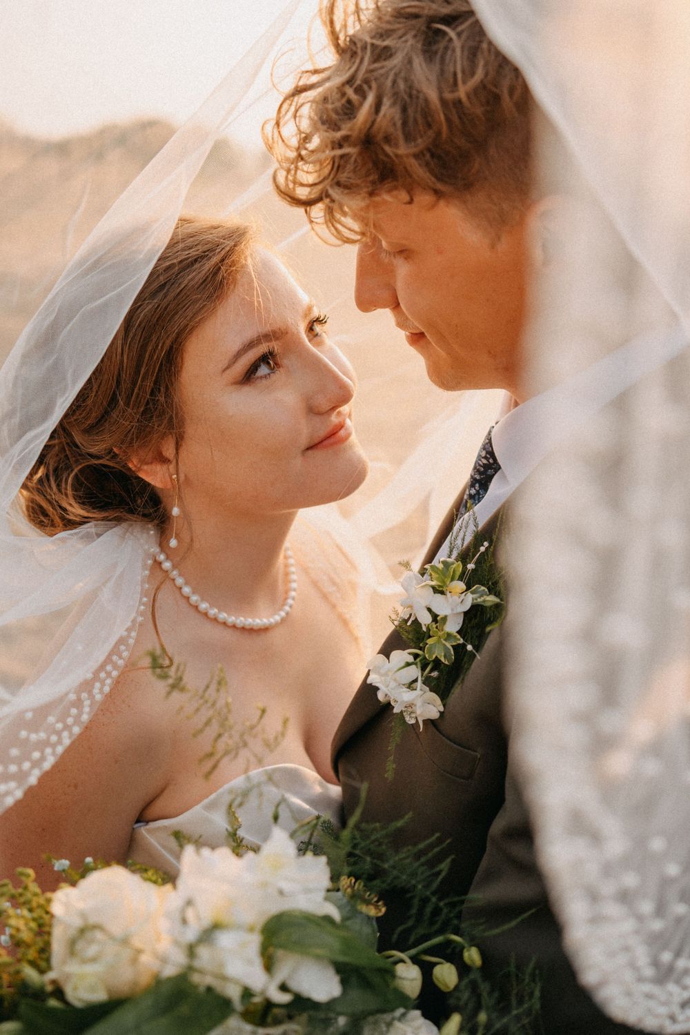 Romantic wedding portrait of a couple in warm sunlight with the bride wearing a veil and pearl necklace.