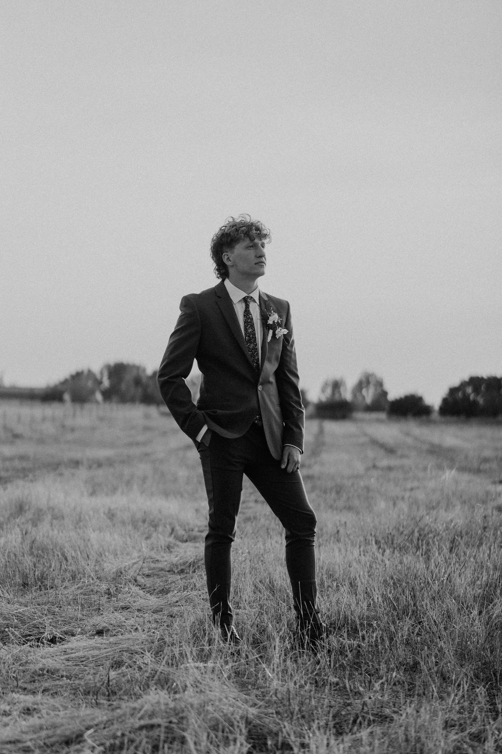 Black and white portraits of groom standing alone in field wearing formal suit.