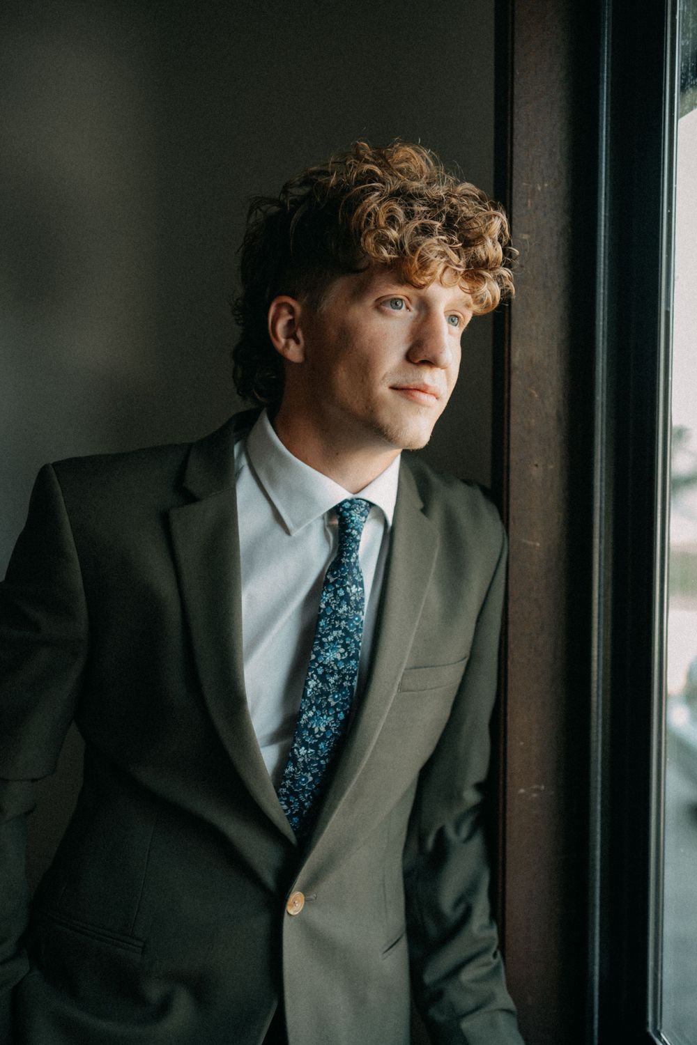 Close-up portrait of a person in a dark suit and patterned blue tie looking pensively out a window.