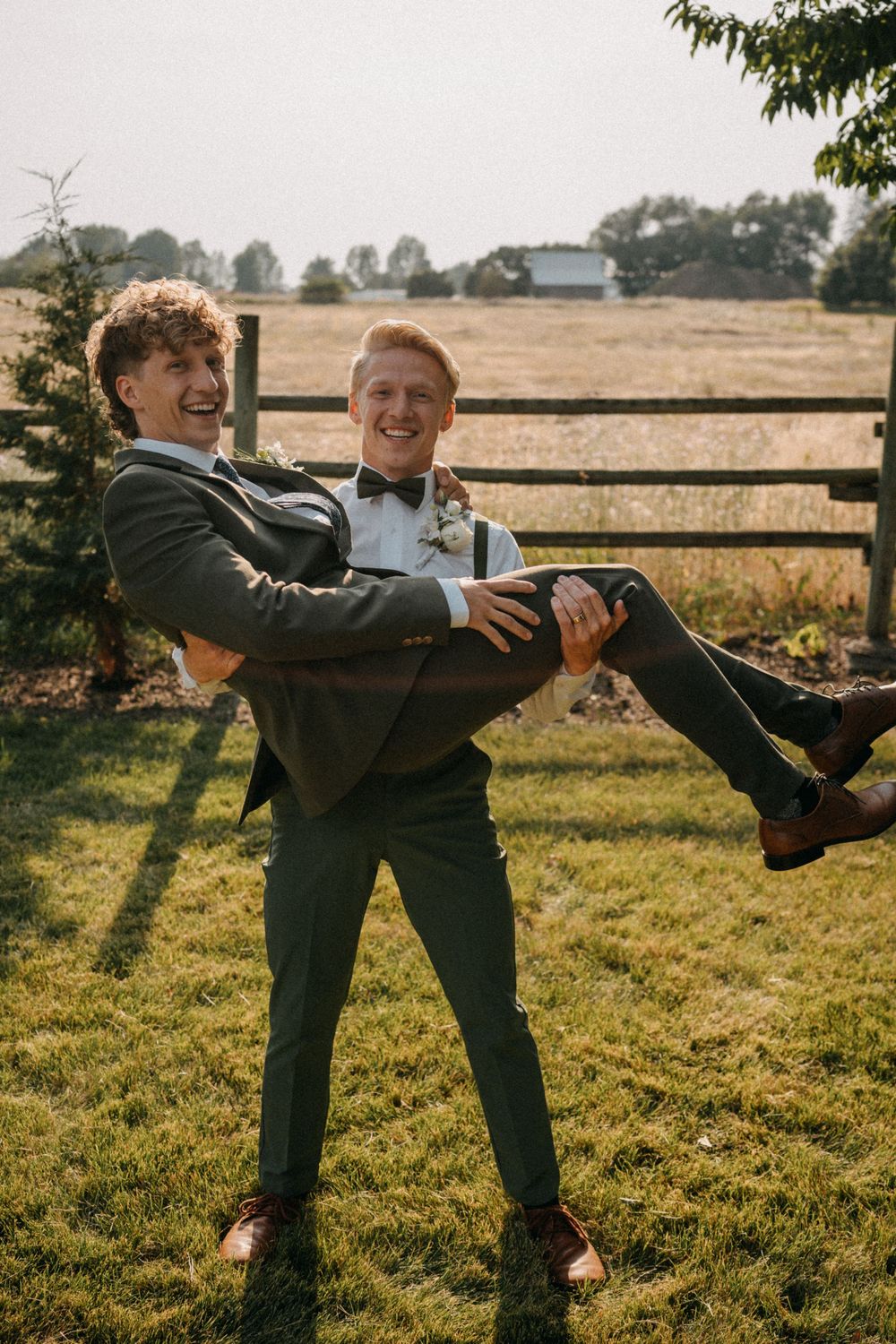 Two people in formal attire playfully posing near a wooden fence at sunset in a rural setting.