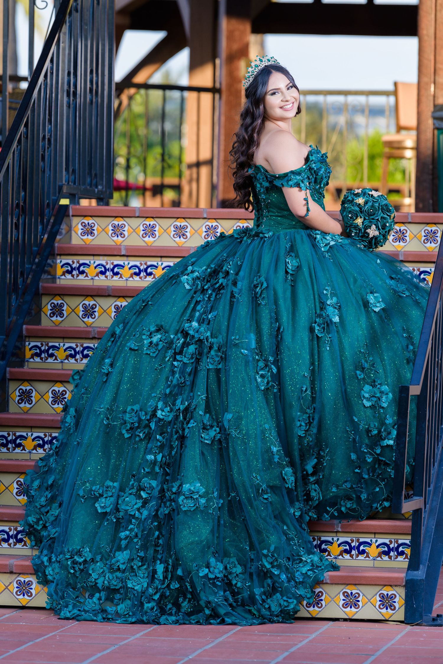 Elegant teal ball gown with floral appliques and off-shoulder design photographed on decorative tiled stairs.