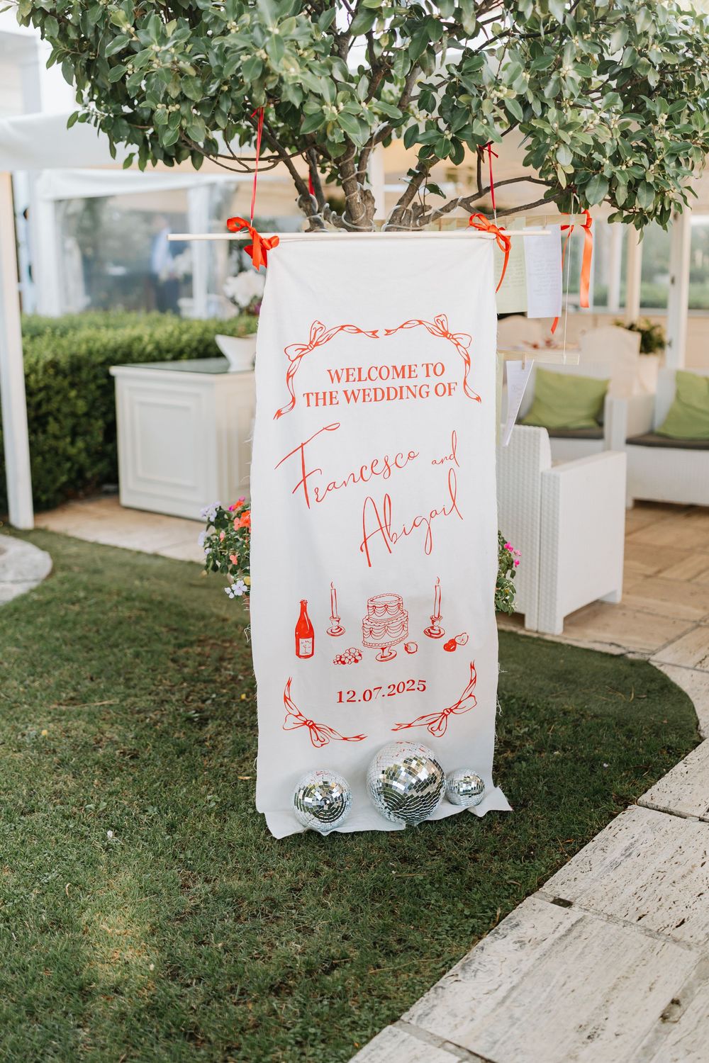 White wedding welcome signs with red calligraphy text displayed on grass and wooden surfaces at an outdoor venue.