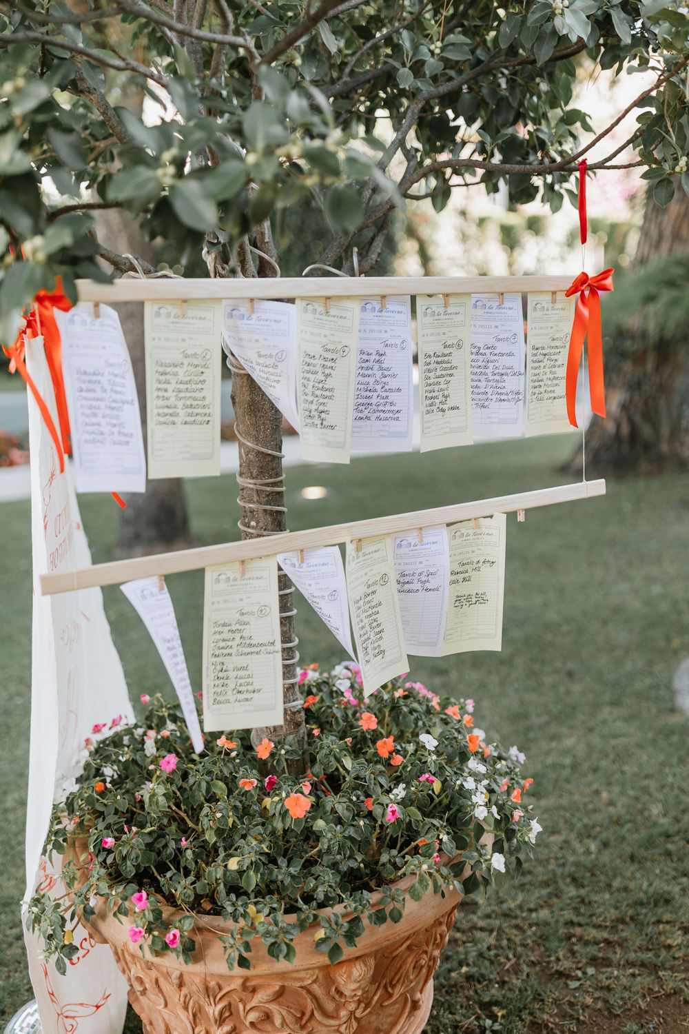 Handwritten seating cards with orange ribbons hang above terracotta planters filled with colorful flowers in an outdoor setting.