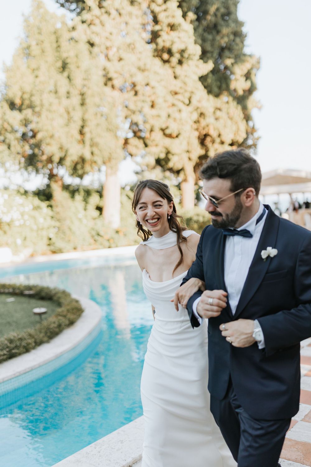 A couple in wedding attire laughs and dances together next to a swimming pool on their special day.