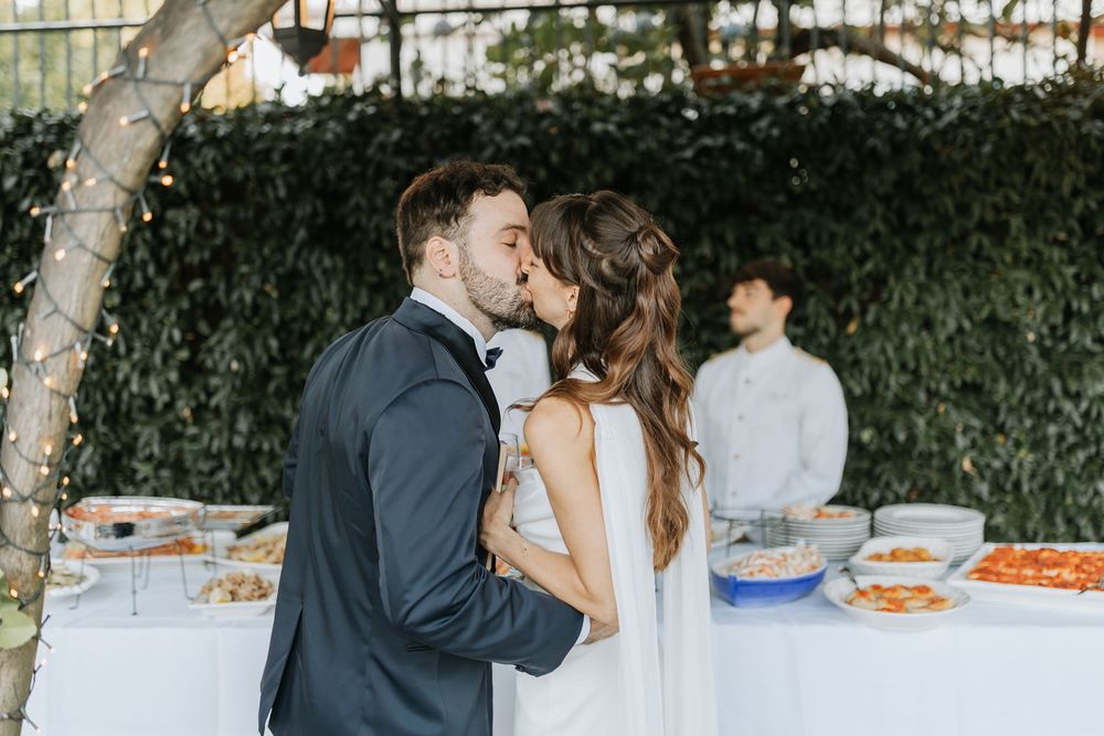 A couple shares a romantic kiss at an outdoor reception buffet with food tables and greenery in the background.