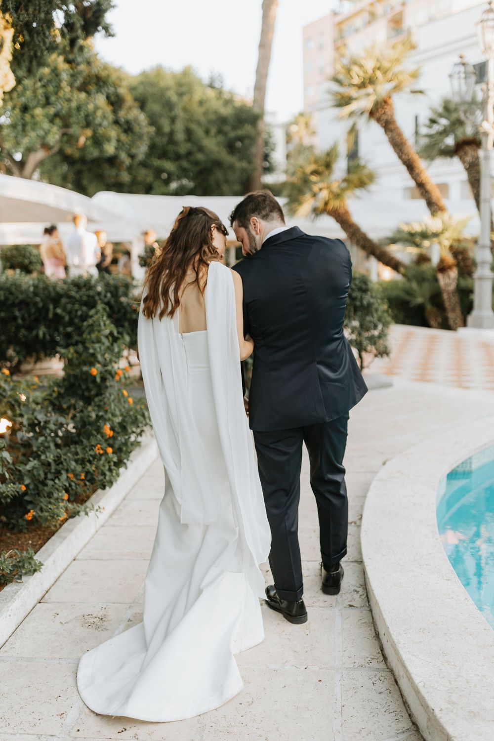 A couple in wedding attire walks along a curved poolside path during a romantic outdoor photoshoot at sunset.