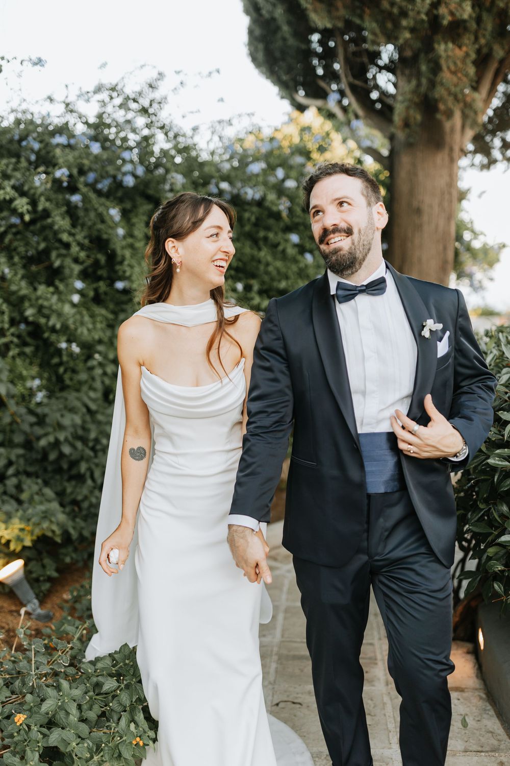 A couple in wedding attire walking together and smiling outdoors with greenery in the background.