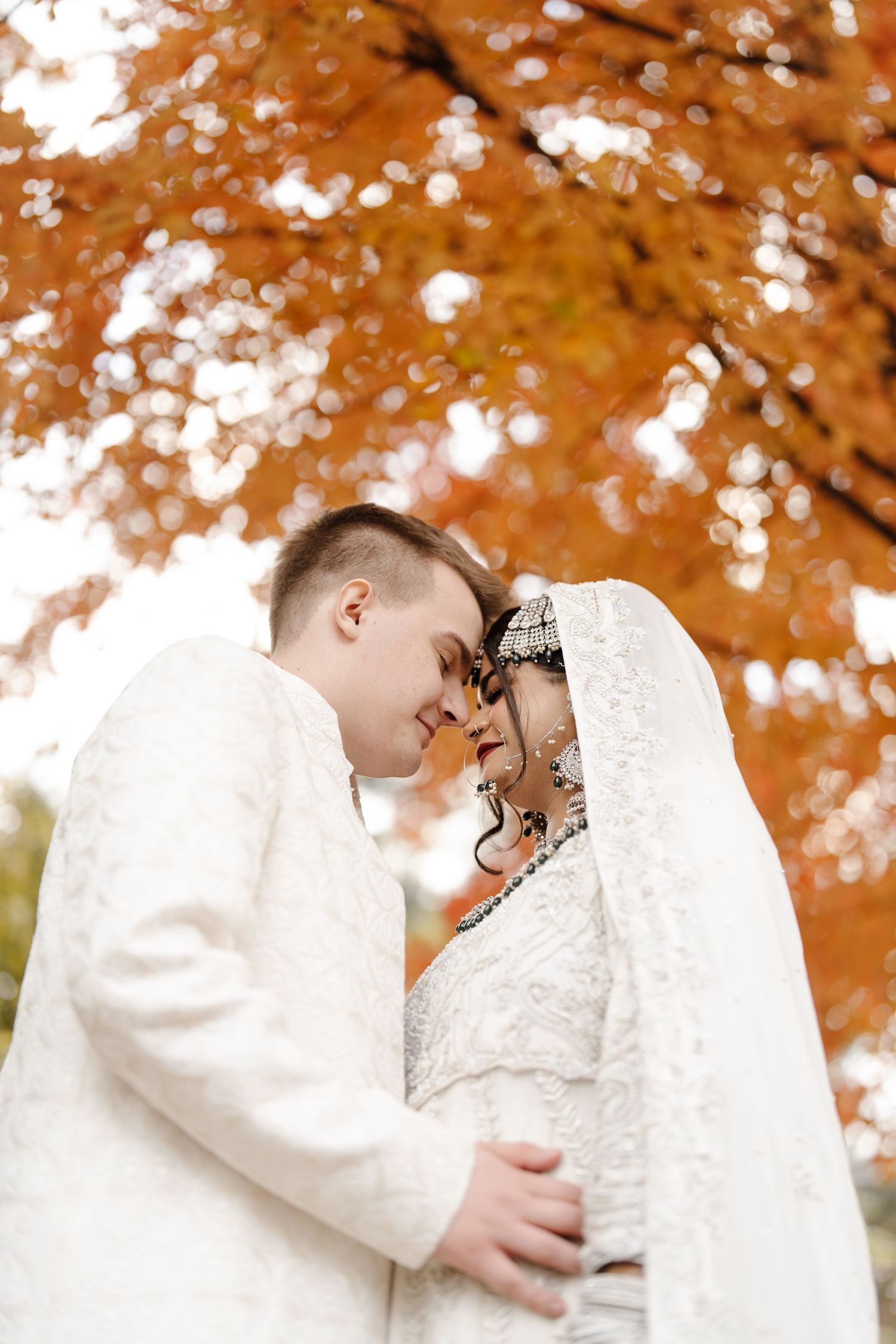 South Asian bride and groom in white traditional attire share intimate moment under vibrant autumn foliage.