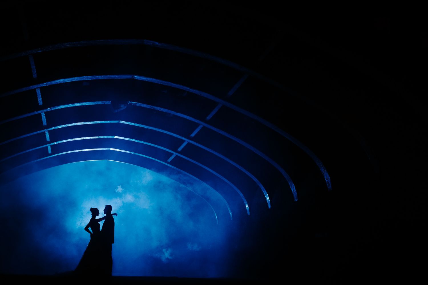 A series of silhouetted figures stand beneath curved architectural structures illuminated by dramatic blue lighting.