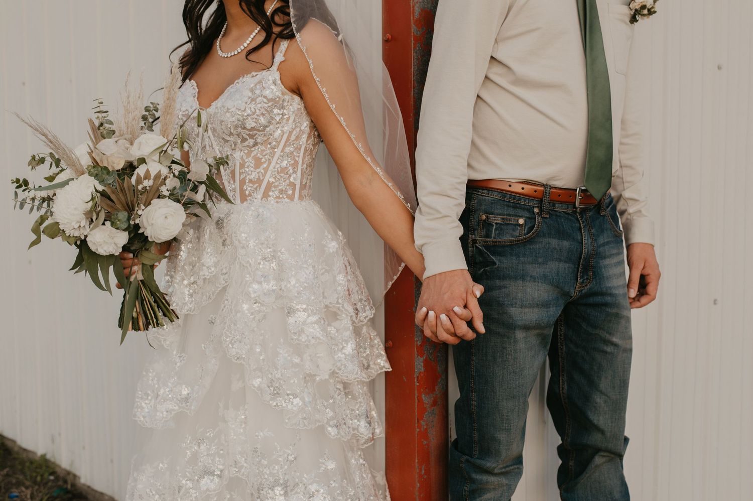 A couple in casual and formal wedding attire holding hands and bouquet against a white wall.