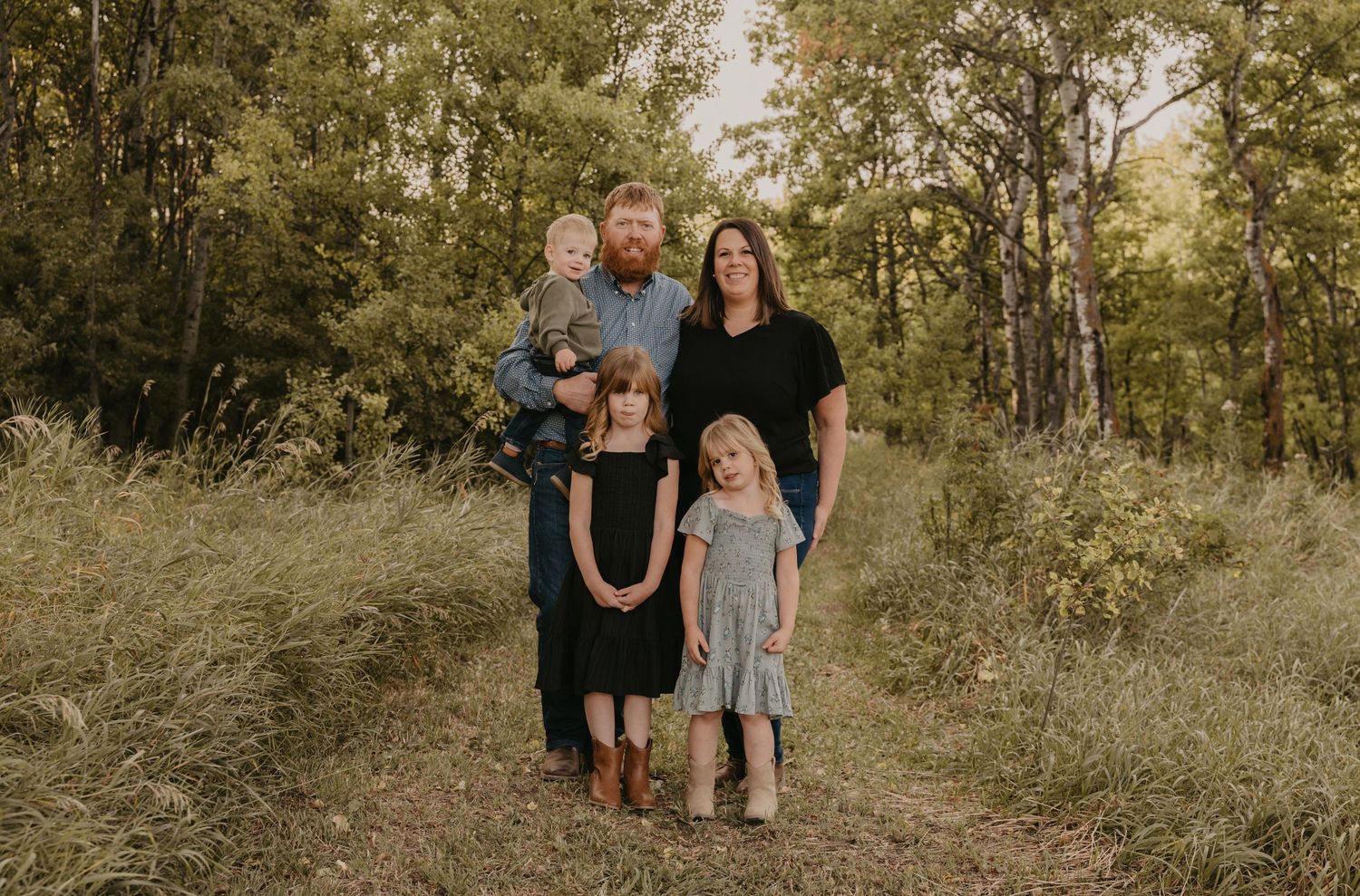 A family stands together on a nature trail surrounded by tall grass and trees during golden hour.