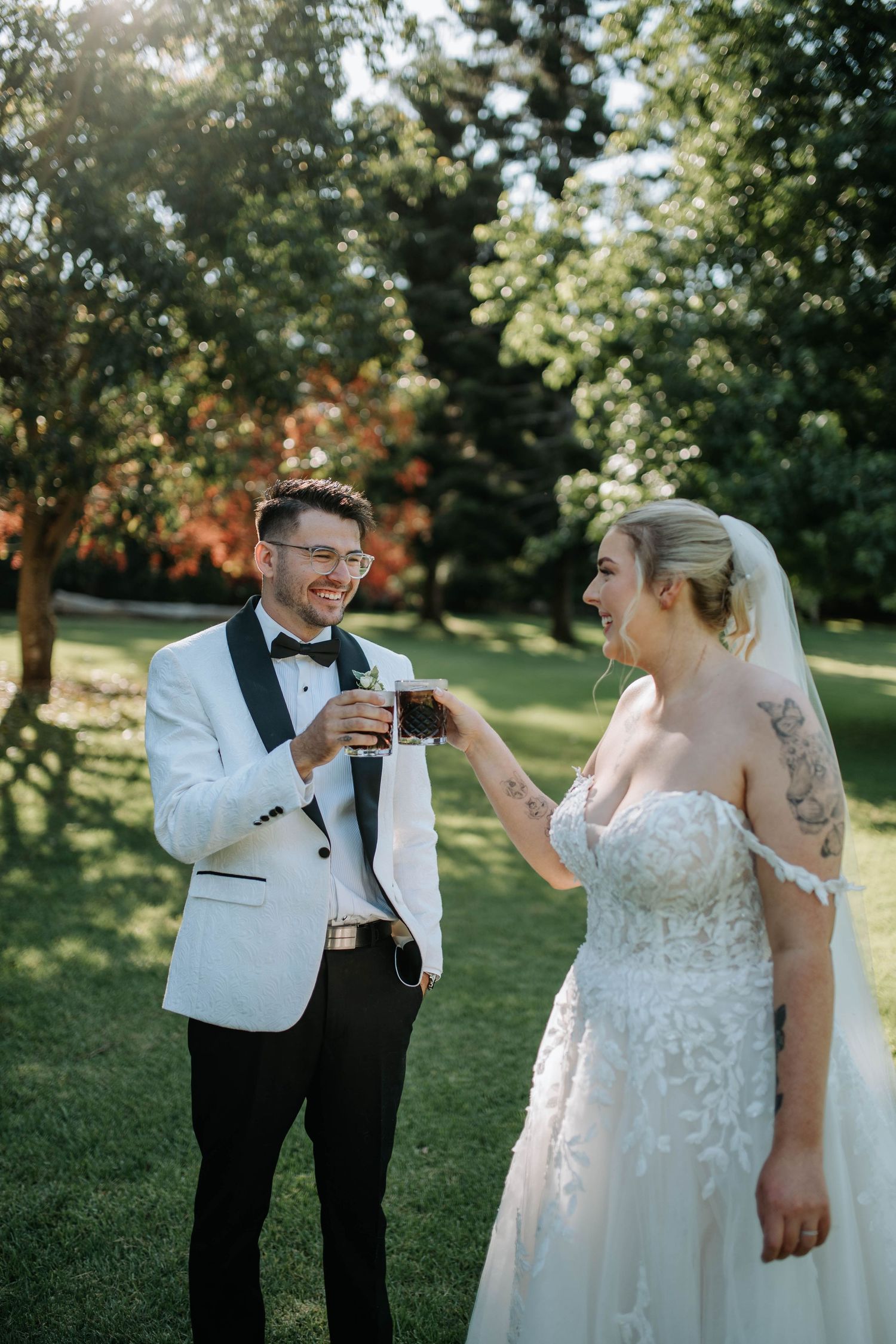 Newlywed couple shares joyful laughter during outdoor wedding photos in a sunlit garden setting.
